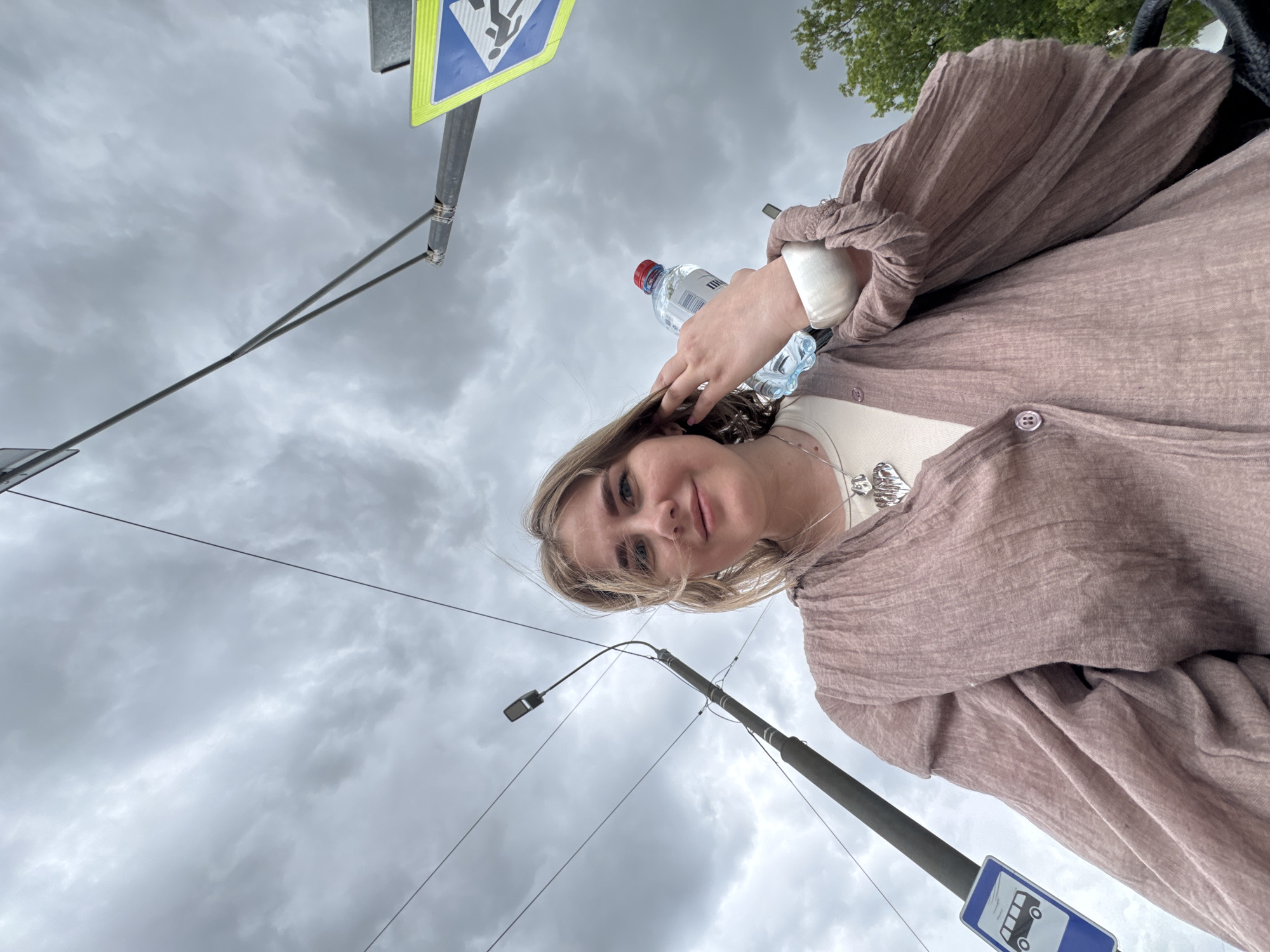 The image captures a young woman in Perm, Russia, likely standing at a bus stop on a cloudy day. She is wearing a light brown blouse and a white undershirt. In her hands, she holds a bottle of water. In the background, there is a bus stop sign and a pedestrian crossing sign, along with a streetlight. The sky is overcast with heavy clouds.