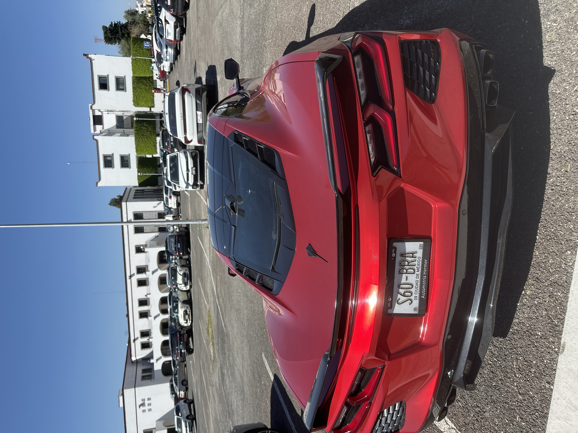 An outdoor, high-angle photograph captures a vibrant red Chevrolet Corvette C8 parked in a large asphalt lot under a clear, bright blue sky. The car, featuring a black roof, engine cover with a black 'Stingray' emblem, and black rear diffuser, is the central focus, positioned diagonally from the bottom right towards the top left. Strong sunlight casts distinct shadows, suggesting a midday or early afternoon setting.
Behind the Corvette, a line of various parked vehicles, including white and dark-colored cars, extends into the background. To the left, several multi-story white buildings with multiple windows and green ivy-covered sections are visible, characteristic of Mexican architecture. More parked cars are lined along these buildings.
Notable details on the Corvette include its distinctive rear taillights, quad exhaust pipes, and the black rear spoiler. The Mexican license plate, partially illuminated by a sun glare, reads "S60-BRA". The license plate frame contains additional text: "09 CIUDAD DE MÉXICO" and "Automotriz Hermer". The setting appears to be a parking area, possibly near commercial or residential buildings, in San Andrés Cholula, Mexico. No people are visible in the image.