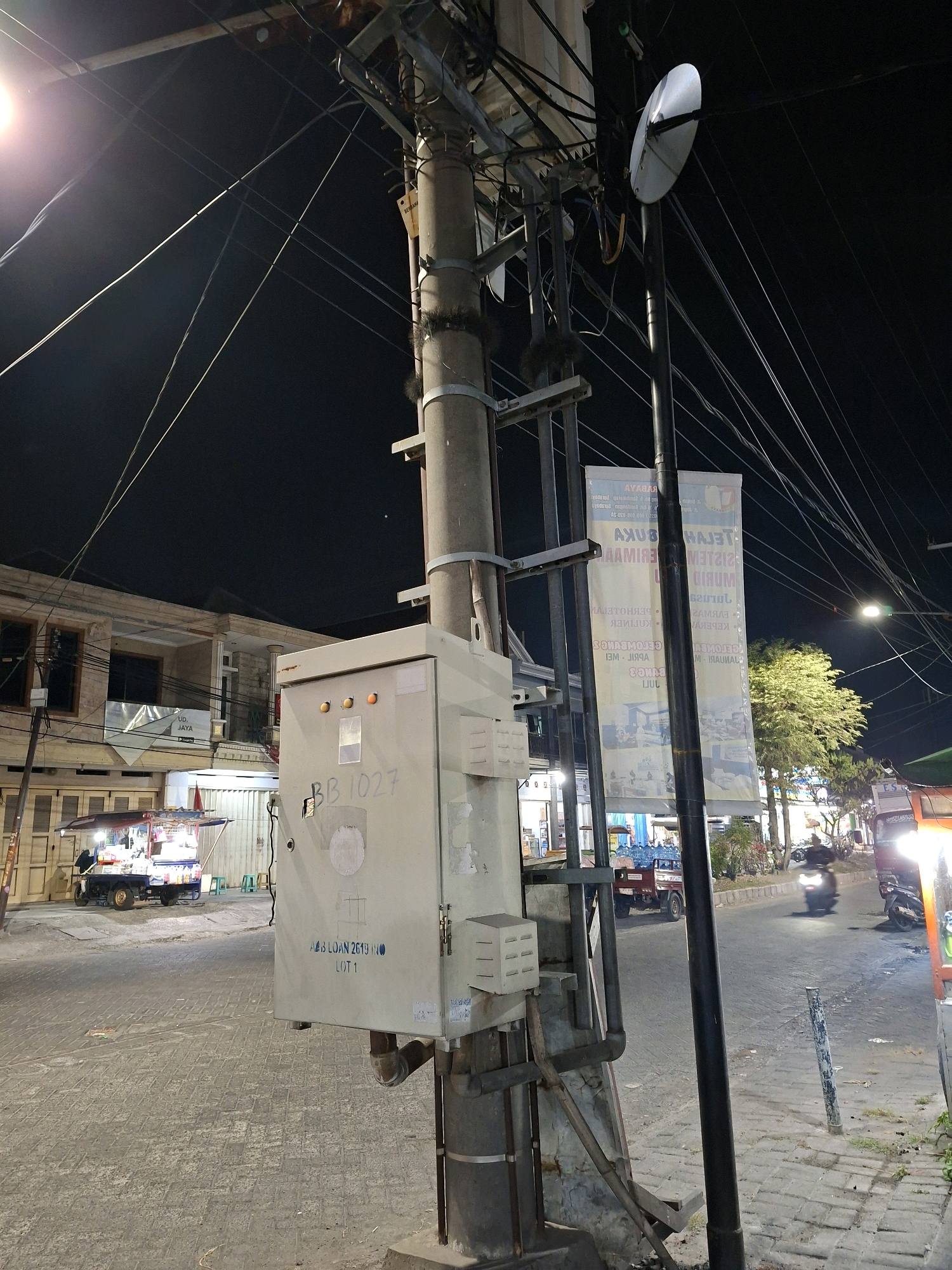 The image depicts a street scene at night in Surabaya, Indonesia. A large, gray electrical utility box is prominently positioned in the foreground, mounted on a concrete pole that is interwoven with numerous black electrical wires. The box has a label that reads "BB 1027" and below that "AS LOAN 2613 (10) LOT 1". To the left of the utility box, a small food stall with brightly lit displays is visible, set up on the side of the road. Behind the stall, a two-story building with several windows and balconies stands. The street is paved with interlocking concrete blocks and appears to be moderately trafficked, with a motorcycle visible in the middle ground to the right, illuminated by streetlights and headlights. A tall, dark pole with a satellite dish attached to its top is also a notable element in the scene, standing to the right of the utility pole. Two large vertical banners, featuring text and images, are attached to the utility pole. The overall environment suggests a commercial or residential street illuminated by artificial lighting.
