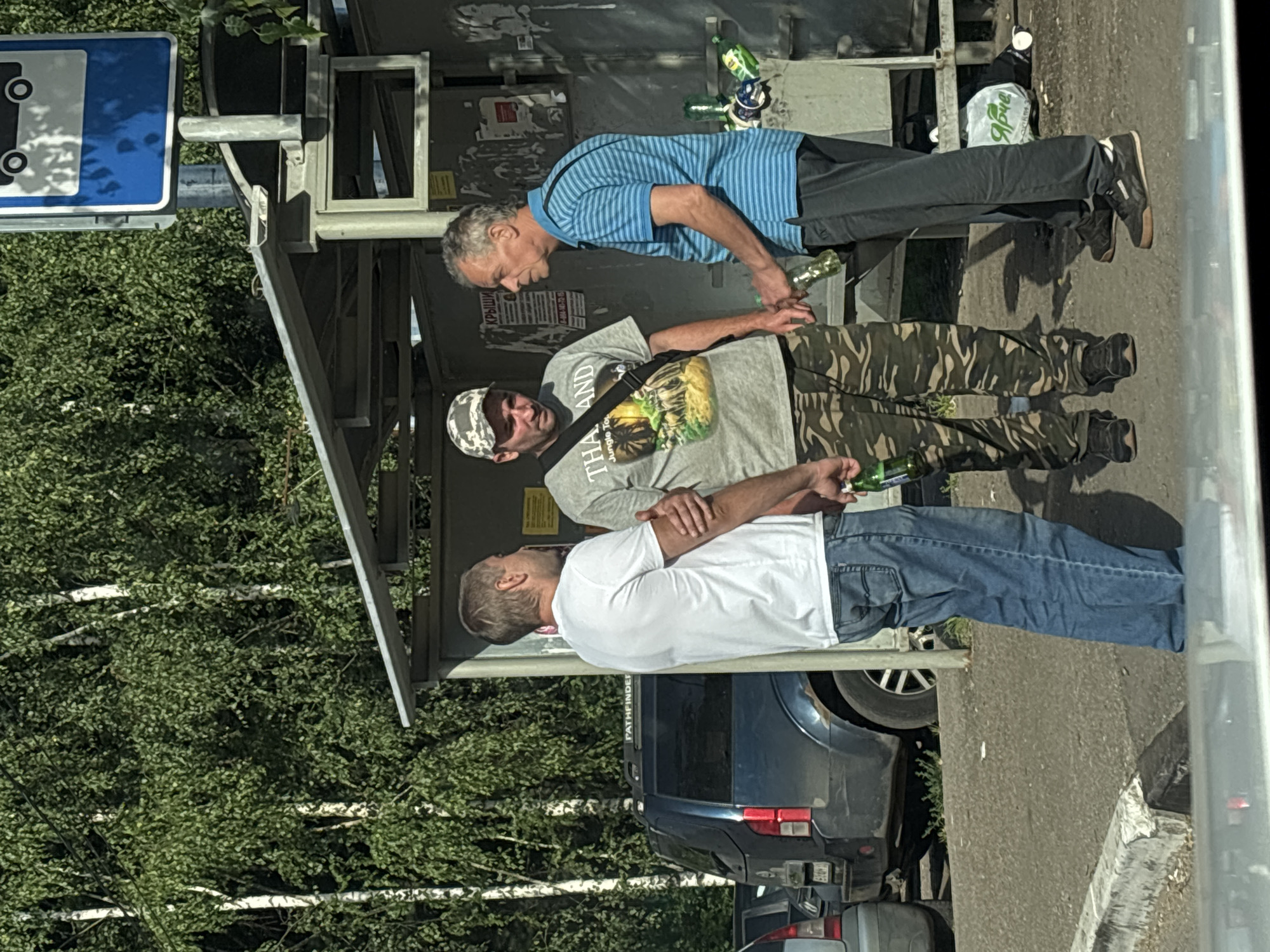 Three men are standing at a bus stop on a sunny day, with trees and a parked SUV in the background. The bus stop has a metal roof and the men appear to be engaged in conversation. One man on the left is wearing a white t-shirt and jeans, holding a green bottle. The middle man is wearing a grey t-shirt with "THAILAND Jungle Tour" printed on it, a camouflage baseball cap, and camouflage pants, also holding a green bottle. The man on the right is wearing a blue and white striped shirt and dark pants, holding a green bottle. Several other green bottles are scattered on the ground near the bus stop. The location is identified as Горбольницы №45, Russia.