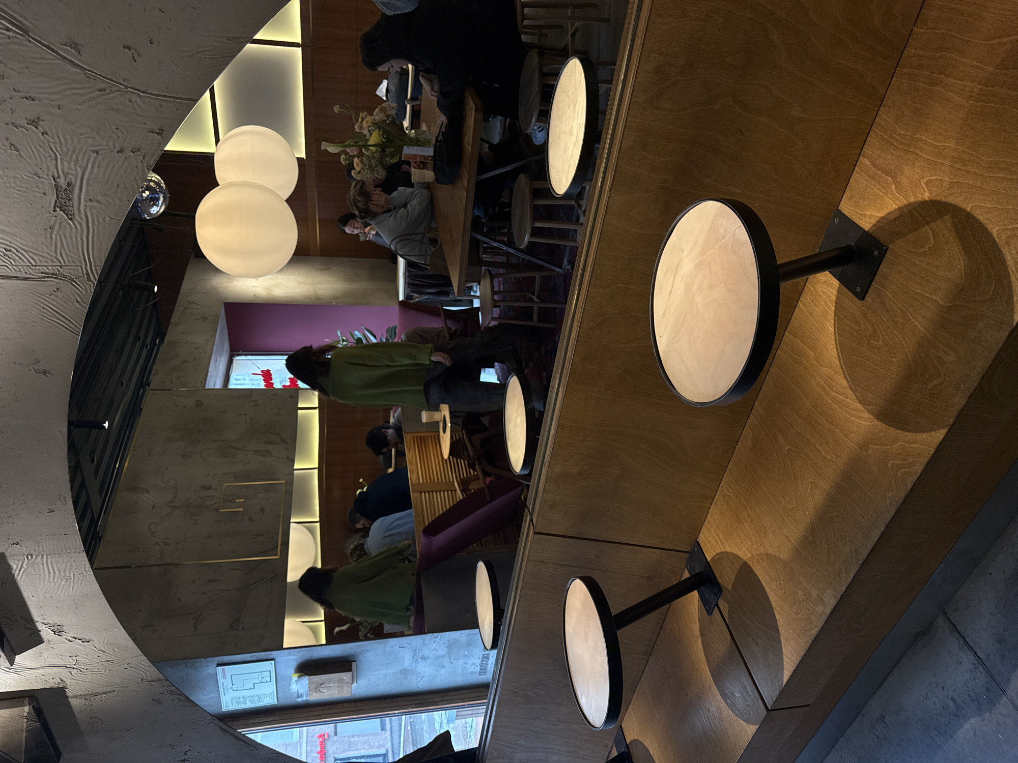 The image, rotated 90 degrees clockwise from its original orientation, captures the interior of a modern cafe or restaurant, likely during the evening. The setting features a contemporary design with a mix of concrete-textured walls, warm wooden paneling, and a long wooden counter or bar that extends diagonally from the foreground to the mid-ground.
Several patrons are visible within the establishment. In the mid-ground, individuals are seated at wooden tables, some appearing to be socializing or looking at their phones. One person in a black hoodie is seated near a table adorned with a floral arrangement. Further back, a person wearing a green top is also seated at a table. People generally appear relaxed and engaged in casual activities.
Lighting is a prominent element of the scene. Two large, spherical white pendant lights hang from the ceiling, providing soft ambient illumination. Along the wooden counter in the foreground, multiple smaller circular light fixtures are mounted on black arms, casting distinct circular shadows on the wooden surface. Integrated strip lighting also illuminates a section of the wall behind the spherical lights. A small disco ball is visible, reflecting light in the upper left area of the image.
Other notable details include a partially visible purple upholstered chair and a window offering a glimpse of an exterior street scene, with some red text visible in reflection. On a wall near the window, a white paper sheet with a diagram, likely a floor plan or safety instruction, is posted, where the Cyrillic word "ПЛАН" (PLAN) can be faintly read. The overall atmosphere conveyed is intimate and stylish.