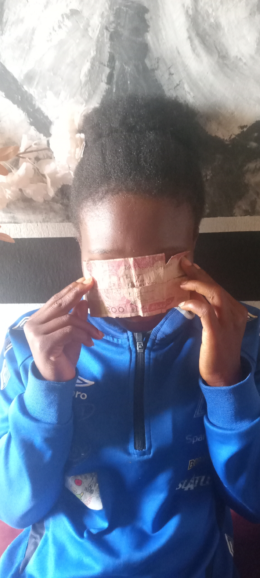 A person is depicted with a two hundred Nigerian Naira note covering their eyes, held in place by their fingers. They are wearing a blue zipper jacket. The background consists of a black and white striped wall, with what appears to be a white floral decoration near the top left. The location and time are not evident from the image.