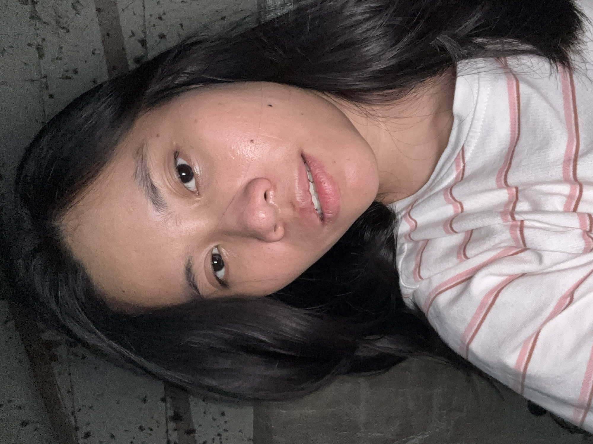 An overhead, medium close-up shot shows a young East Asian woman looking into the camera with her head tilted to the left. She has long, dark, wavy hair that falls around her face. She is wearing a white t-shirt with thin pink and brown stripes. The background appears to be a textured, dark gray wall. This image was taken in East Jakarta, Indonesia.