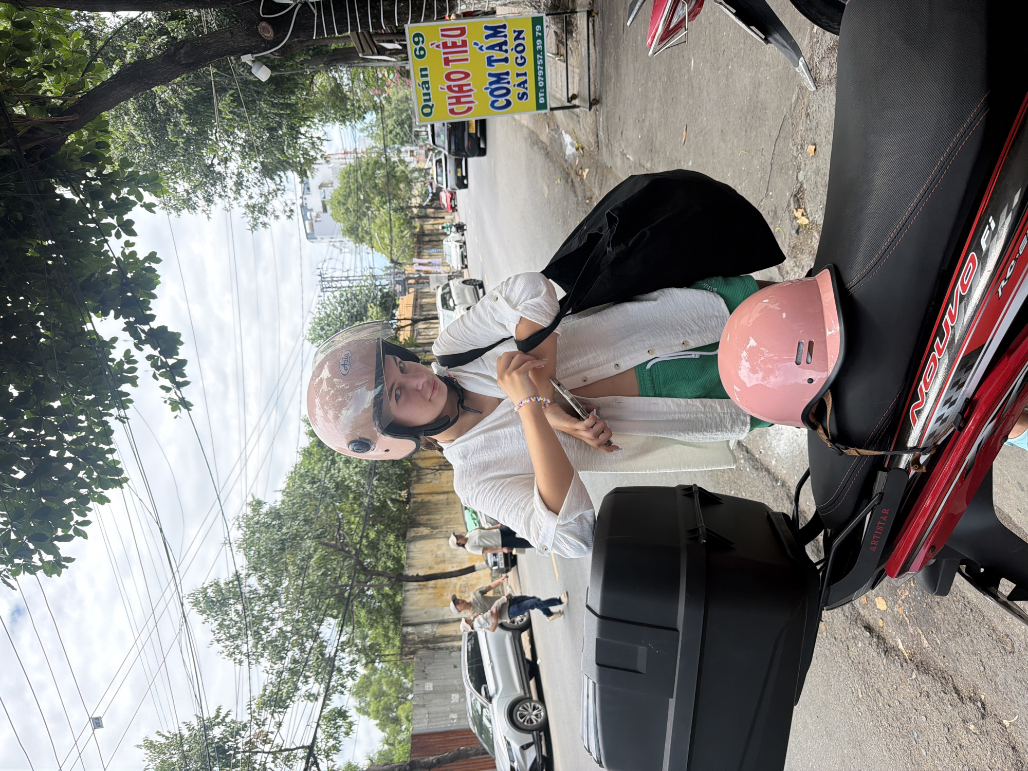 In Nha Trang, Vietnam, a young woman wearing a light pink helmet and a white, open button-down shirt stands next to a parked motorcycle. Her green shorts are visible beneath the shirt. She holds her left hand to her chest, appearing to adjust something on her shirt, while her right hand holds a mobile phone and a bracelet with colorful beads adorns her wrist. A black tote bag is slung over her left shoulder.
In the foreground, the motorcycle's black seat is prominently displayed, with a pink helmet resting on it. A black cargo box is mounted on the rear of the motorcycle. The background reveals a street scene with a sign in Vietnamese indicating "Quán 69 Cháo Tiều Cơm Tấm Sài Gòn" and a phone number. Cars and other motorcycles are visible on the street, and trees line the sidewalk. The sky is overcast. A man carrying a child walks across the street in the mid-ground, with another person walking nearby. The scene is captured outdoors in a city environment.