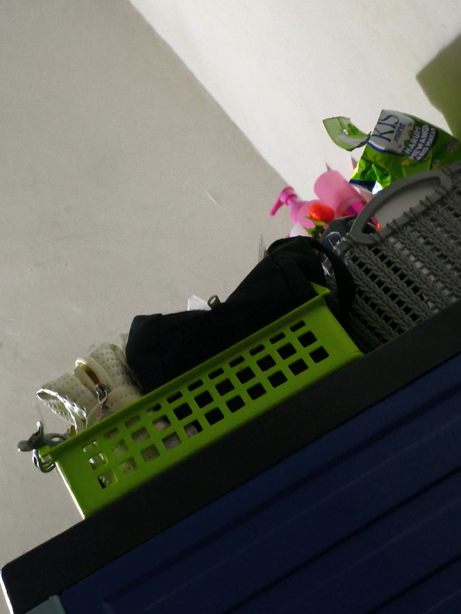 The image shows a low-angle, close-up shot of items on a shelf within a residence in Karangtalun, Indonesia. In the foreground, a bright green plastic basket with a grid pattern is positioned on top of a dark blue surface, likely a piece of furniture such as a cabinet or dresser. Inside the green basket, several off-white items, possibly slippers or shoes, are visible, along with a silver clip attached to the basket's edge. Behind the green basket, a black fabric item, potentially a bag or piece of clothing, is present. Further back and to the right, a gray plastic basket with a similar grid pattern is partially visible, containing items such as a pink spray bottle and a green packet of "KIS mint" candies. The background consists of a plain, light-colored wall, with a portion of a ceiling visible at the top of the frame. The lighting suggests an indoor environment, possibly with natural light entering from the side.