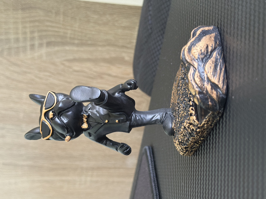 A matte black figurine of a stylized French bulldog, accented with metallic gold details, is positioned on a dark, horizontally ribbed surface, likely a desk mat. The figurine wears a black suit jacket with gold buttons, a gold bow tie, and black trousers. It features gold-framed sunglasses and a light-colored cigar protruding from the right side of its mouth. The figure stands on its left leg, with its right leg bent at the knee and raised, presenting the sole of the foot forward. It rests on an oblong, textured base, colored black with metallic gold highlights, which includes a horizontal, rectangular depression at its front designed for supporting an object. The background is out of focus, showing a light brown, vertically oriented wood grain pattern, consistent with a wall panel or furniture, with a darker, indistinct object visible to the right. The scene is located in Sofia, Bulgaria.