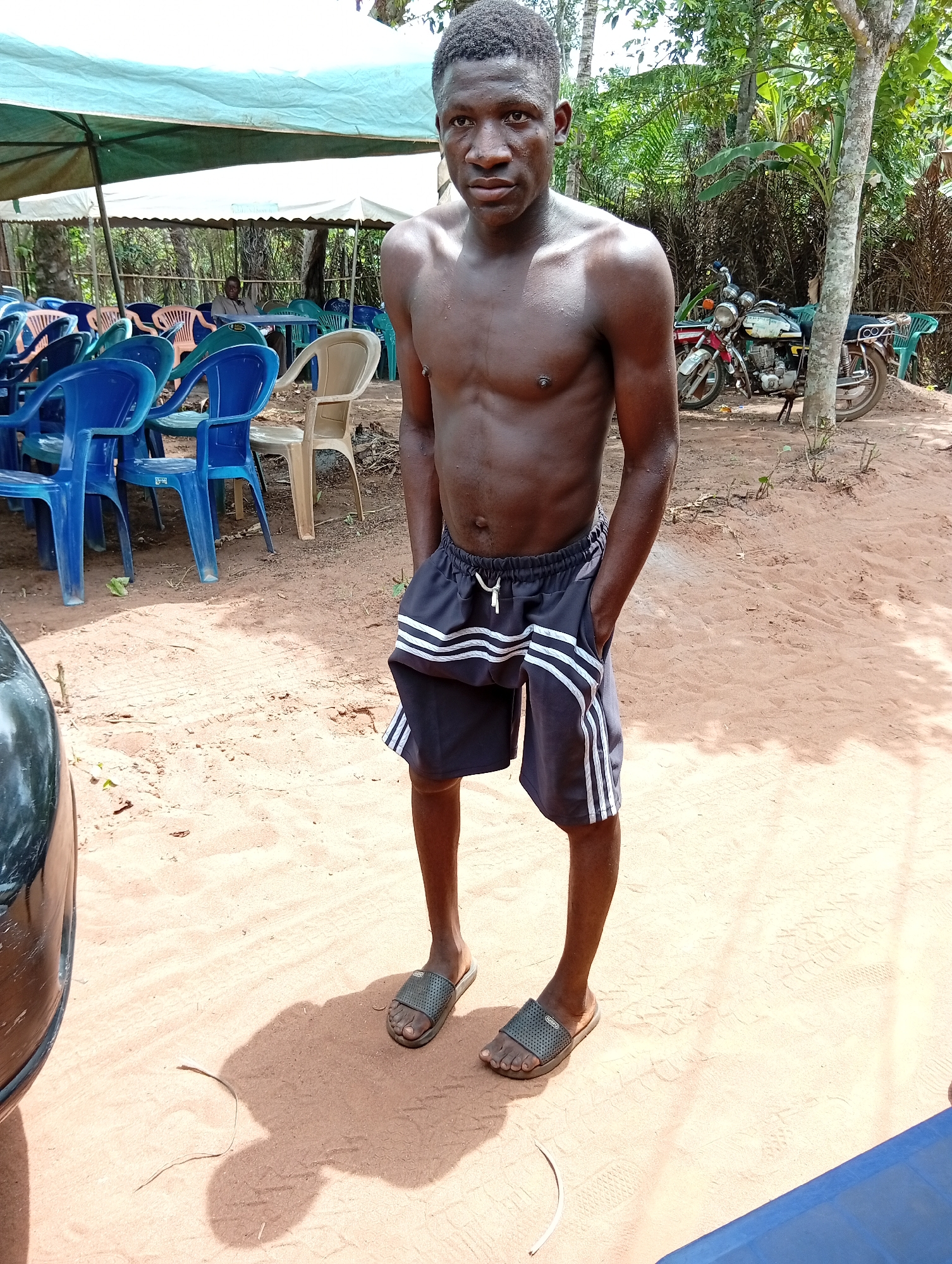 A young Black man stands shirtless on reddish-brown sandy ground, looking directly at the camera with a neutral expression. He wears dark athletic shorts with white stripes on the sides and dark slide sandals, with his hands in his pockets. His skin appears slightly shiny, suggesting warmth or recent activity.
The setting is an outdoor gathering area in Umundugba, Nigeria, characterized by bright sunlight casting strong shadows, indicating midday. In the background, several light blue and white canopies provide shade over numerous plastic chairs in blue, light green, and beige, arranged in rows. A person is visible seated at a table under one of the distant canopies, partially obscured by the chairs.
To the right, against a backdrop of lush tropical trees and foliage, two motorcycles are parked, one red and white, the other green and white. Part of a dark, possibly metallic, object or vehicle is visible in the lower left foreground. The scene conveys a casual, outdoor social or event space in a warm, tropical climate. No discernible text is visible.