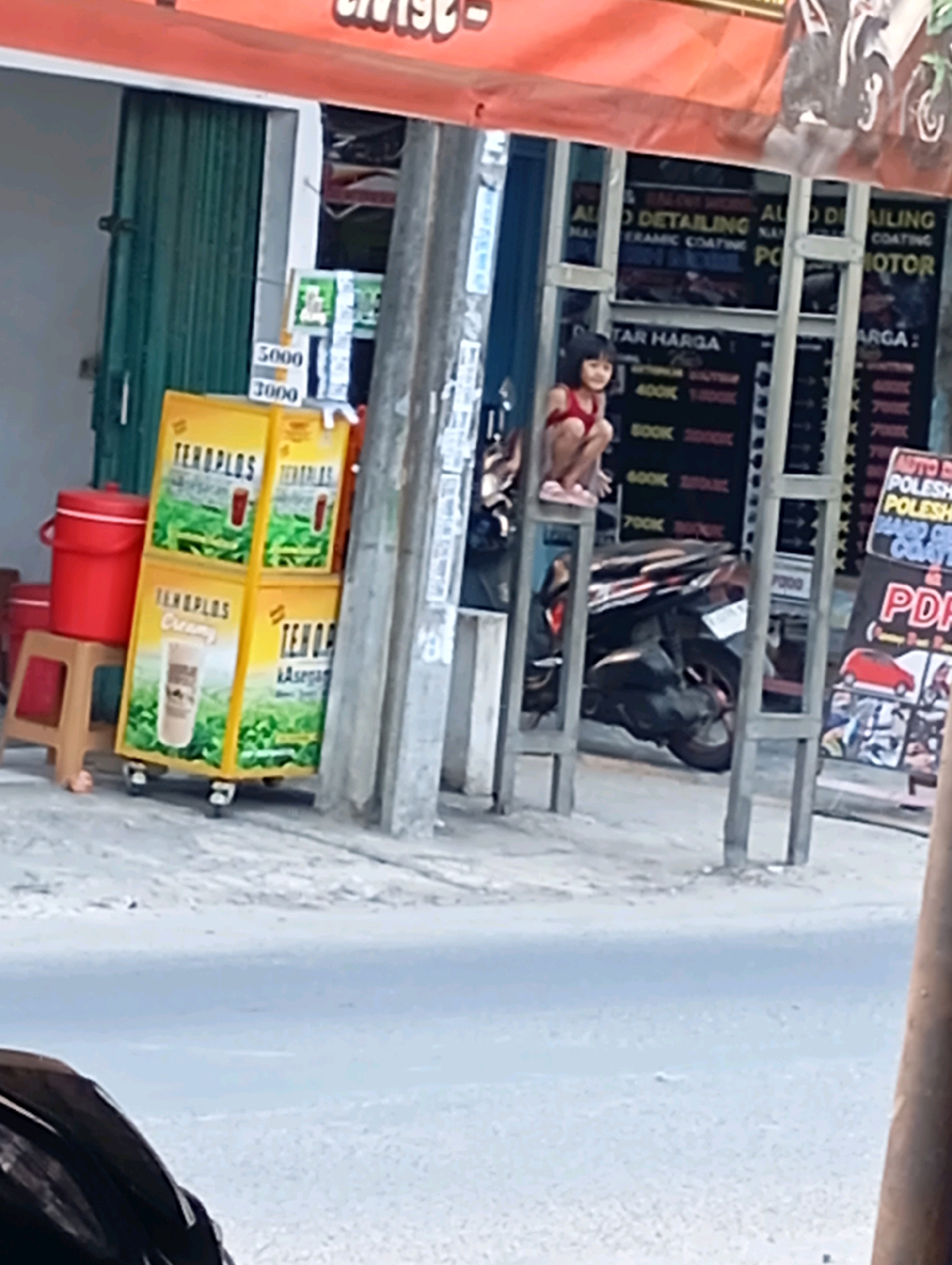 In Babelan, Indonesia, a young girl in a red top and shorts sits on a metal frame structure, appearing relaxed. She is the primary subject, with a backdrop of a storefront and street scene. To her left, stacks of yellow boxes labeled "TENOPLOS" are visible, suggesting a beverage or food product. One box displays the word "Creamy" and another "Asega," accompanied by images of drinks. A red cooler and a small stool are also present nearby.
Behind the girl and the product display, a dark-colored motorcycle is parked. The storefront itself features signs with text in English, advertising services like "AUTO DETAILING," "CERAMIC COATING," and "POLISH." Prices are listed alongside some services, such as "400K," "500K," and "700K." A prominent sign reads "PDF." The scene appears to be set outdoors during daylight, possibly in the morning or afternoon, given the lighting. The overall impression is that of a casual moment captured in a commercial area.