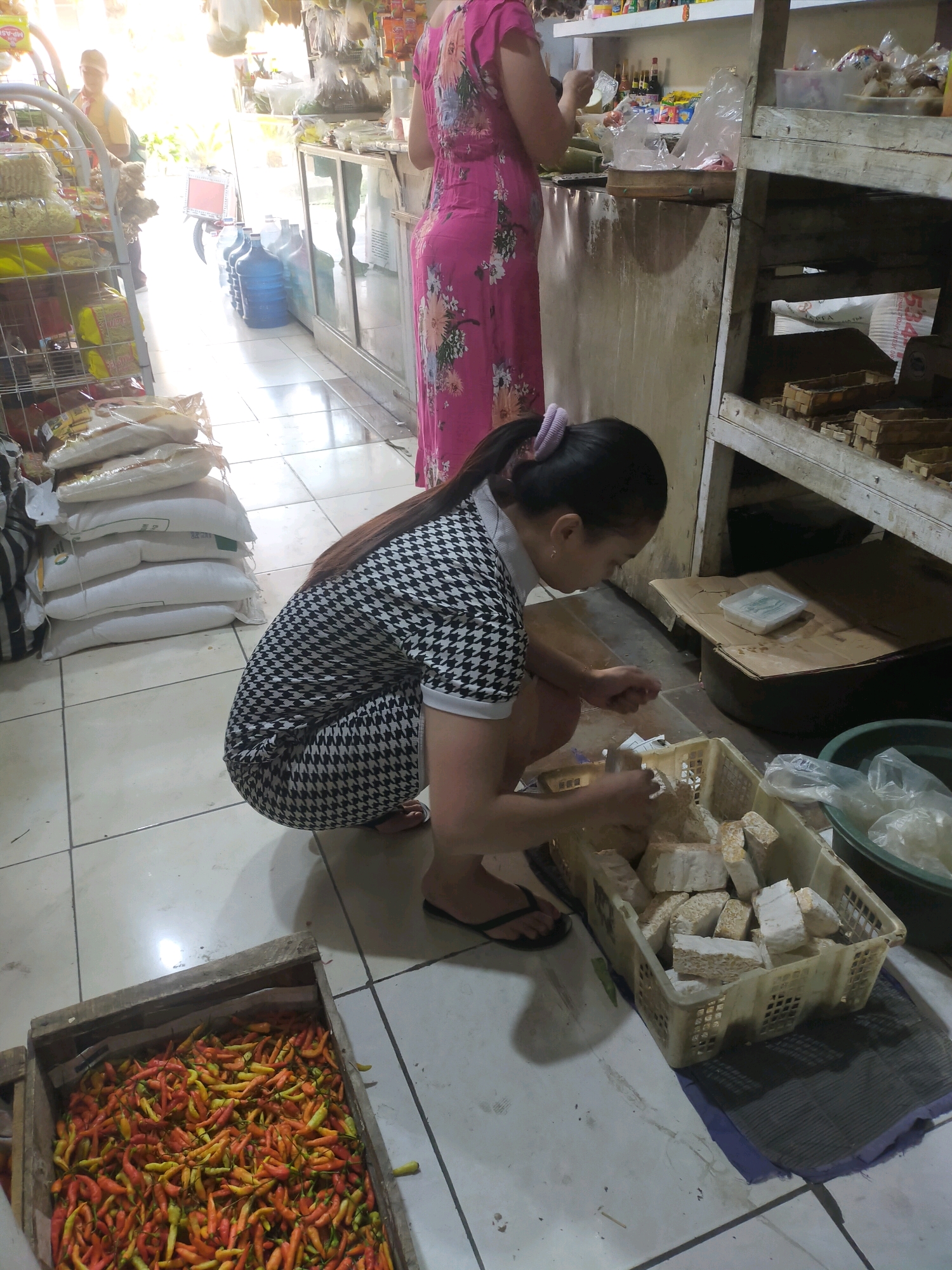 In Mojodanu, Indonesia, inside a brightly lit indoor market stall, a woman with dark hair tied in a ponytail, wearing a black and white houndstooth dress, is crouched down. She is sorting through a white plastic crate filled with rectangular blocks of tempeh, a fermented soybean food. Her hands are actively engaged with the tempeh, suggesting she is either preparing it for sale or packaging it. To her left, a large wooden crate overflows with a vibrant mix of small, colorful chilies, predominantly red, orange, and yellow. In the background, shelves are stocked with various goods, and a woman in a long pink floral dress stands behind a counter. The floor is tiled, and the overall impression is of a busy, functional marketplace.
