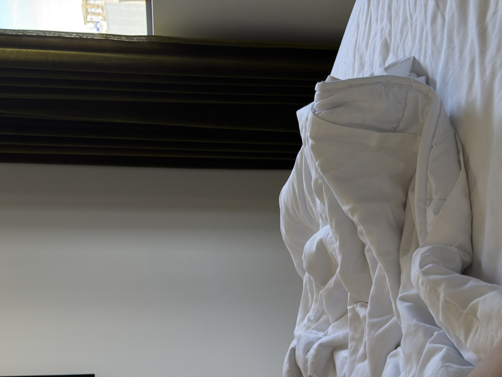 A close-up, low-angle shot depicts a messy white bed with crumpled sheets in what appears to be a hotel room. The bed occupies most of the frame, with its texture and folds creating a sense of disarray. To the right, a dark green velvet curtain is partially drawn, revealing a sliver of a window. Through the window, a bright sky and a building with windows are visible, suggesting an urban setting. A small portion of a person's arm or leg is visible in the lower left corner of the image, partially obscured by the bedsheets. The wall on the left is plain and light-colored. The scene suggests the aftermath of someone having recently occupied the bed.