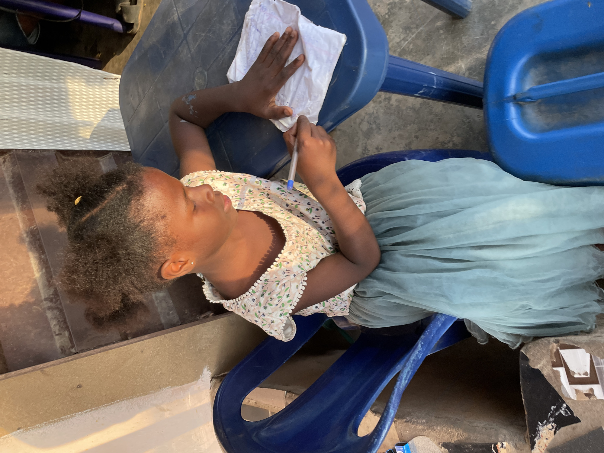 A young girl is sitting on a blue plastic chair at a blue plastic table, writing on a piece of paper with a blue pen. She is wearing a light-colored dress with a floral pattern. The setting appears to be outdoors, possibly in a compound, with a rough concrete floor and some discarded materials in the background. The sunlight suggests it is daytime. The girl's focus is on her writing, with her head tilted down.