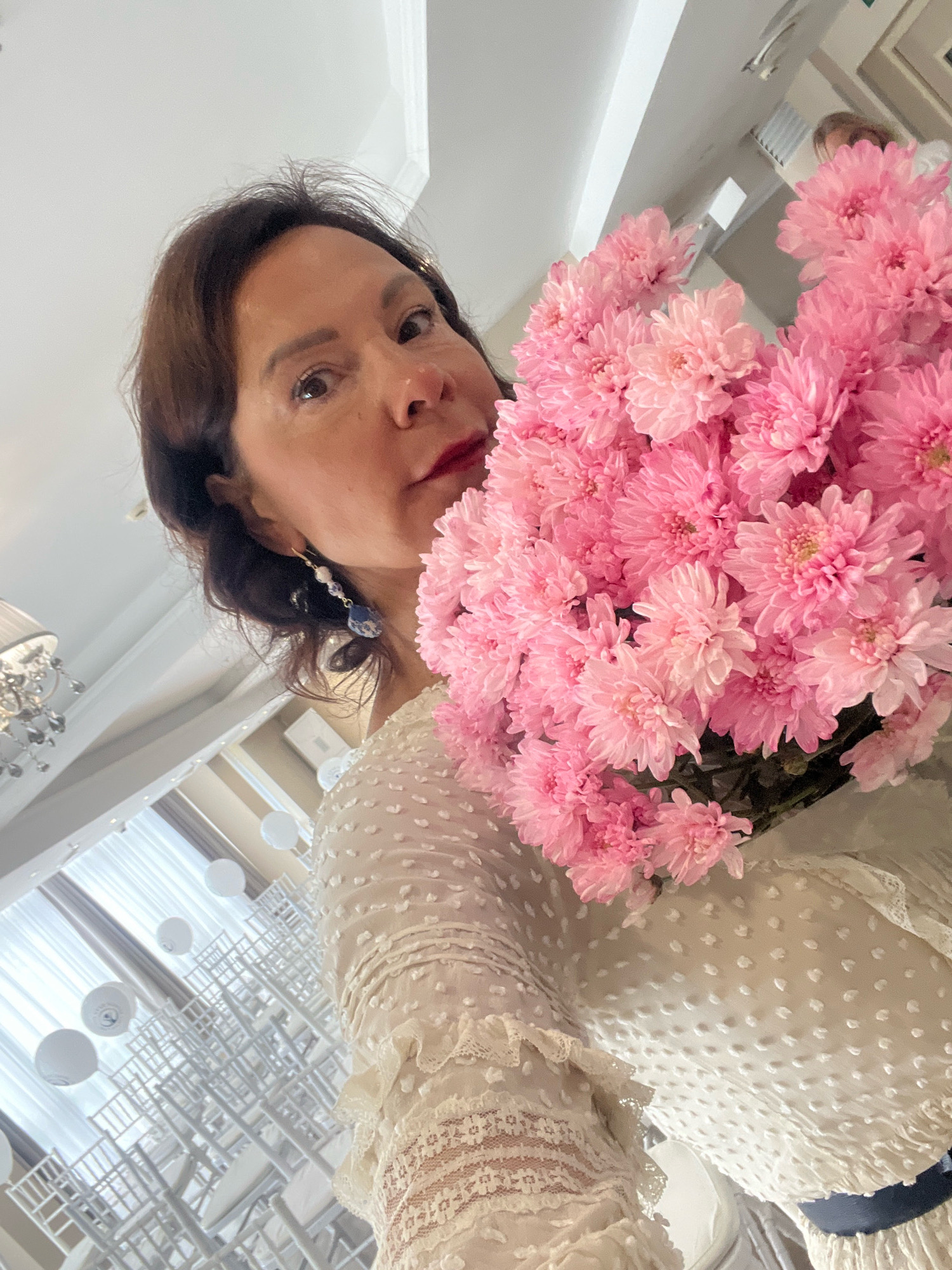 In Sochi, Russia, a woman with dark hair and red lipstick poses for a selfie, holding a bouquet of pink chrysanthemums. She is wearing a white lace dress with intricate detailing. In the background, rows of white chairs are arranged, suggesting a venue for an event. White balloons are also visible, floating near the ceiling. A chandelier hangs from the ceiling, adding to the ambiance of the location. The overall impression is of a formal setting, possibly a wedding or a party.