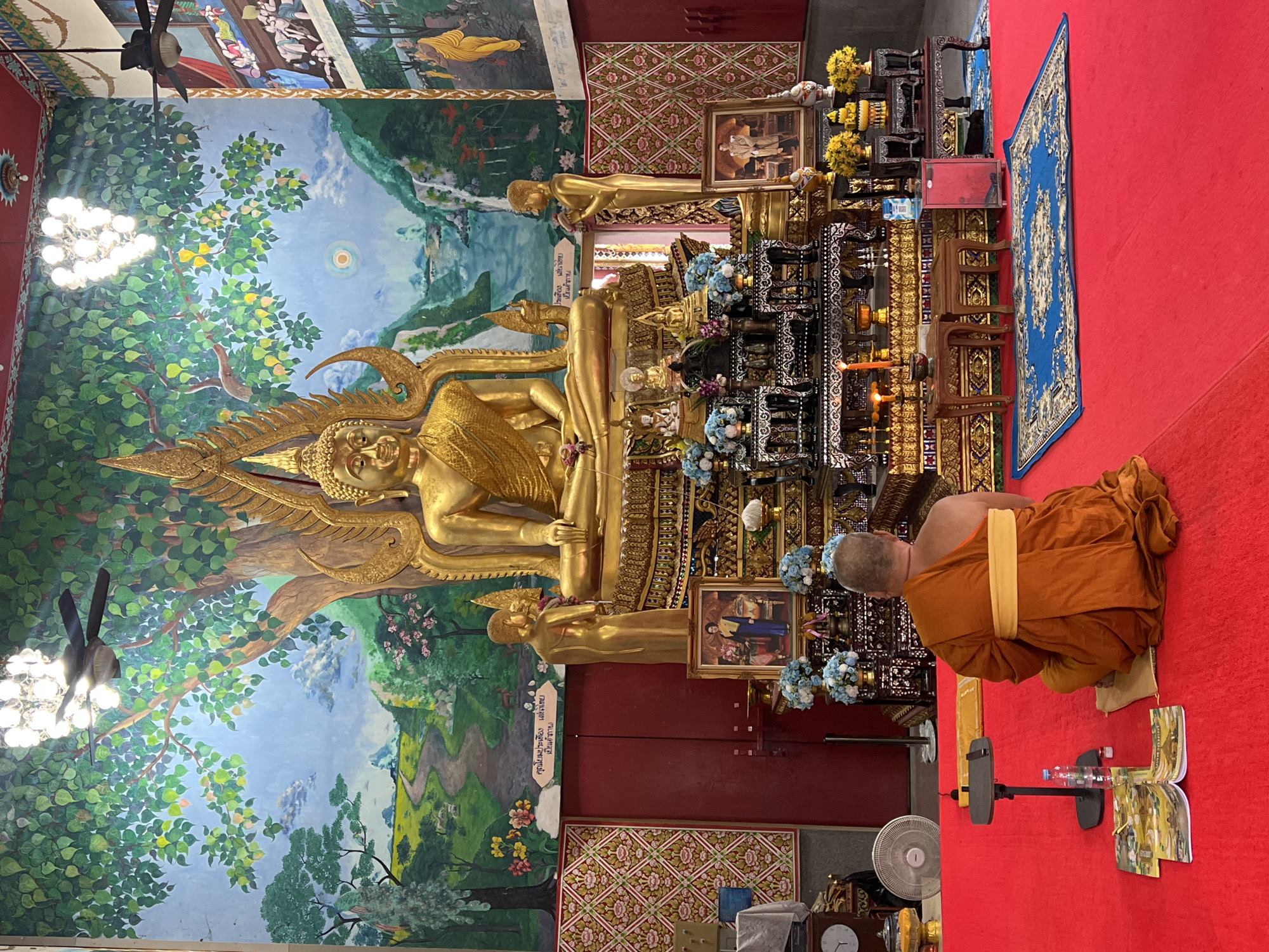 In the interior of a temple in Baan Plai Laem, Thailand, a Buddhist monk in an orange robe sits in meditation on a red carpet. He faces a large, ornate golden Buddha statue. The Buddha statue is positioned in front of a mural depicting a lush landscape with trees, mountains, a river, and a path. To the sides of the Buddha are smaller golden statues and elaborate altar decorations with flowers and offerings. In front of the monk, on the red carpet, are two open books and a bottle of water. The environment includes a ceiling fan, light fixtures, and patterned wall coverings.