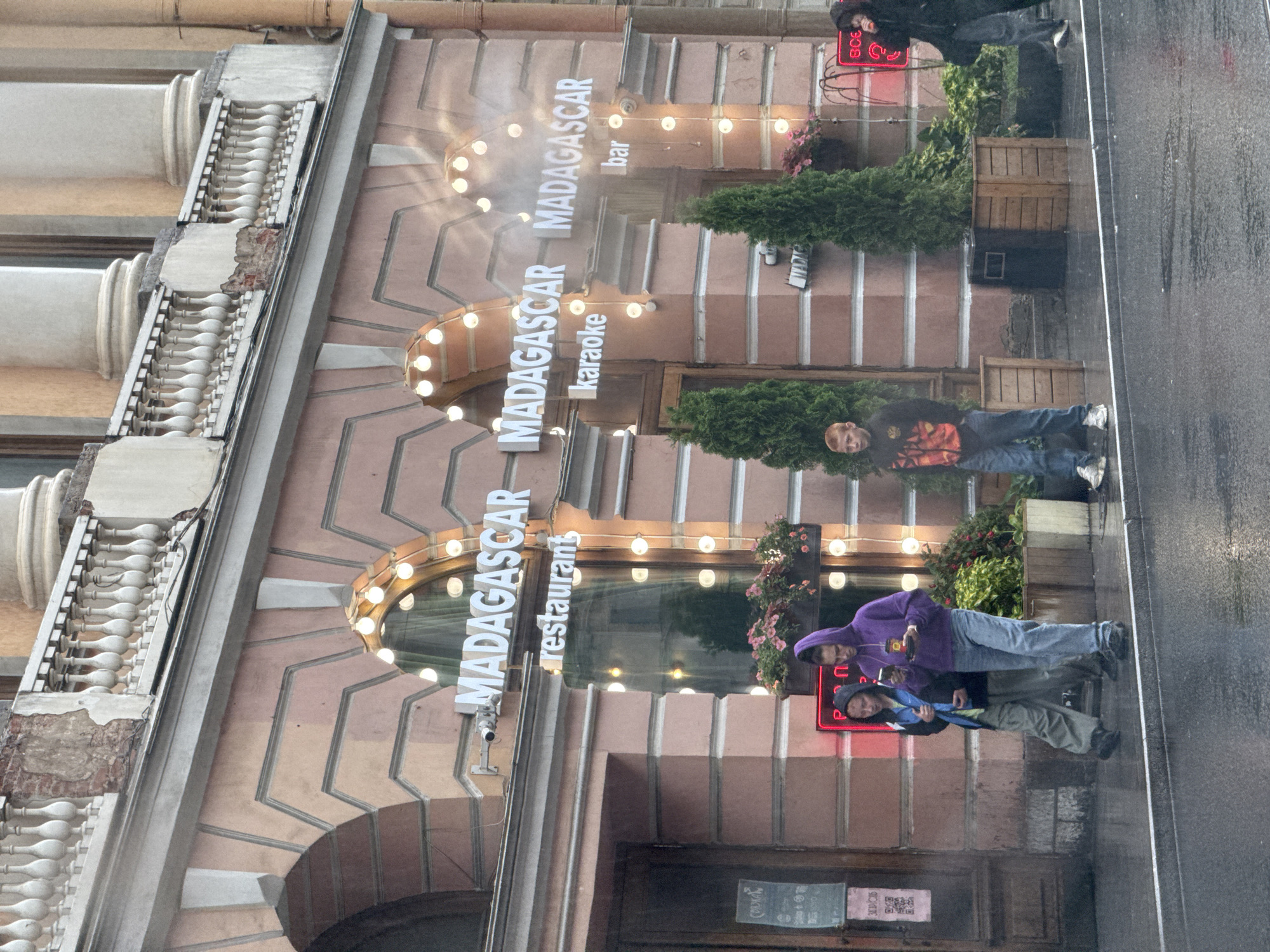 The image shows the facade of a building with signage for "MADAGASCAR restaurant karaoke bar". The building features classical architectural elements, including columns and a balustrade. The facade is painted a pale pinkish-orange and has decorative arched entrances. String lights with exposed bulbs are strung across the facade, illuminating the signage and the storefronts.
In front of the building, there is a wet sidewalk and street, indicating recent or ongoing rain. Several people are visible:
* Two individuals, one wearing a purple hooded sweatshirt and jeans, and the other in a dark jacket and light-colored pants, are walking on the sidewalk. The person in the purple hoodie is looking at their phone.
* A young male with red hair, wearing a black jacket with orange and black detailing and blue jeans, stands on the sidewalk, facing towards the building.
* Another individual, mostly obscured by darkness, is walking away from the building on the right side of the frame.
Green potted plants and trees are placed in front of the building. There are also signs on the doors, one of which appears to be a QR code. The overall atmosphere suggests a European city street scene, likely in Saint Petersburg, Russia, given the architectural style and the presence of Cyrillic text on some signage (though the main signage is in English).