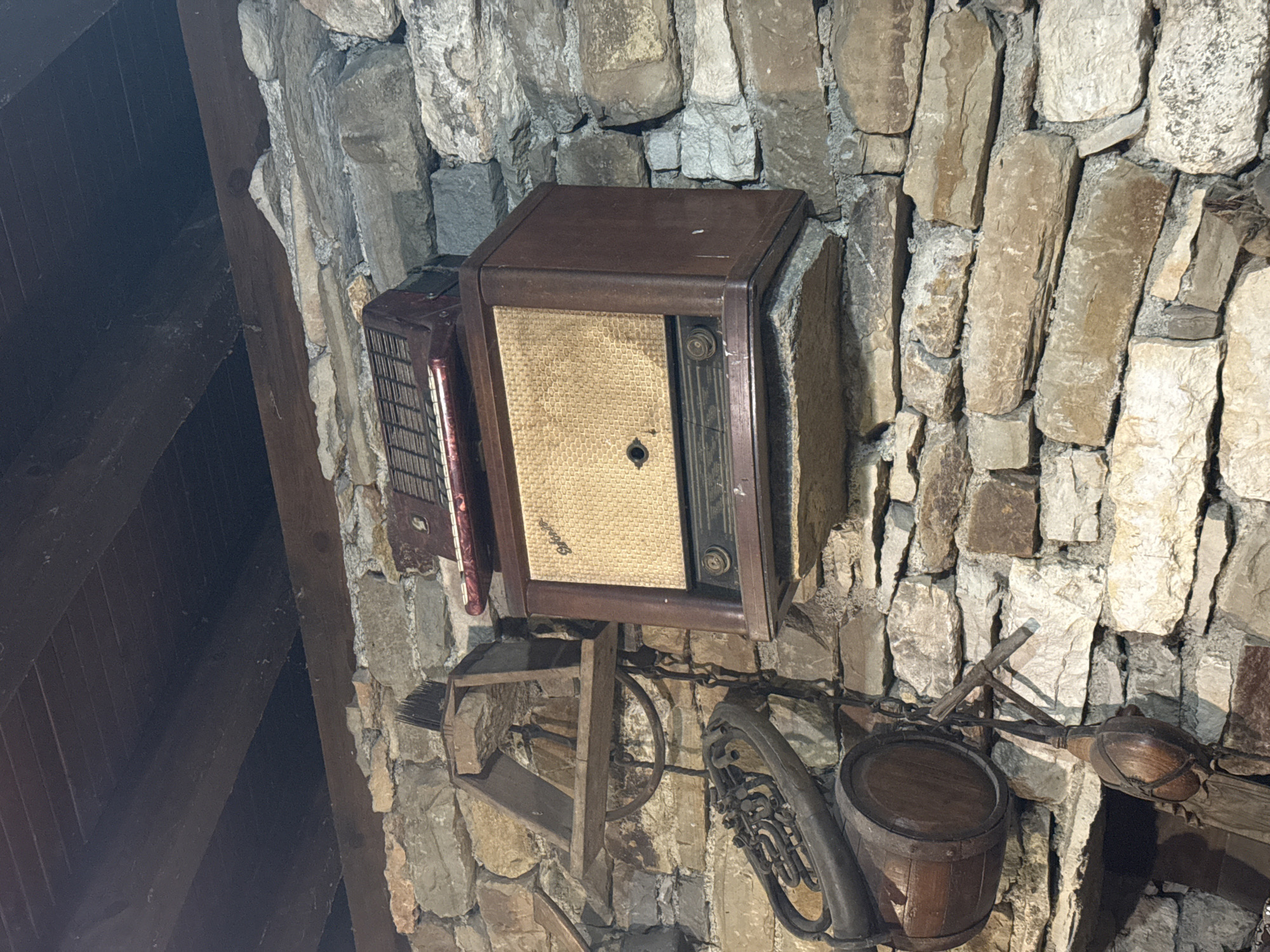 The image depicts a collection of vintage items mounted on a rough stone wall, likely in an interior setting. The objects include a brown wooden radio with a gold speaker grill, an accordion with red keys, a brass-colored musical instrument resembling a sousaphone, a small wooden barrel, and other assorted wooden and metal items hanging by chains. The wall is constructed from irregular, light-colored stones. The ceiling above is made of dark wooden planks. The overall impression is one of rustic decoration or display. The image is an interior shot and does not show any people or outdoor elements. The location of Petrevene, Bulgaria is not visually evident in the image itself.