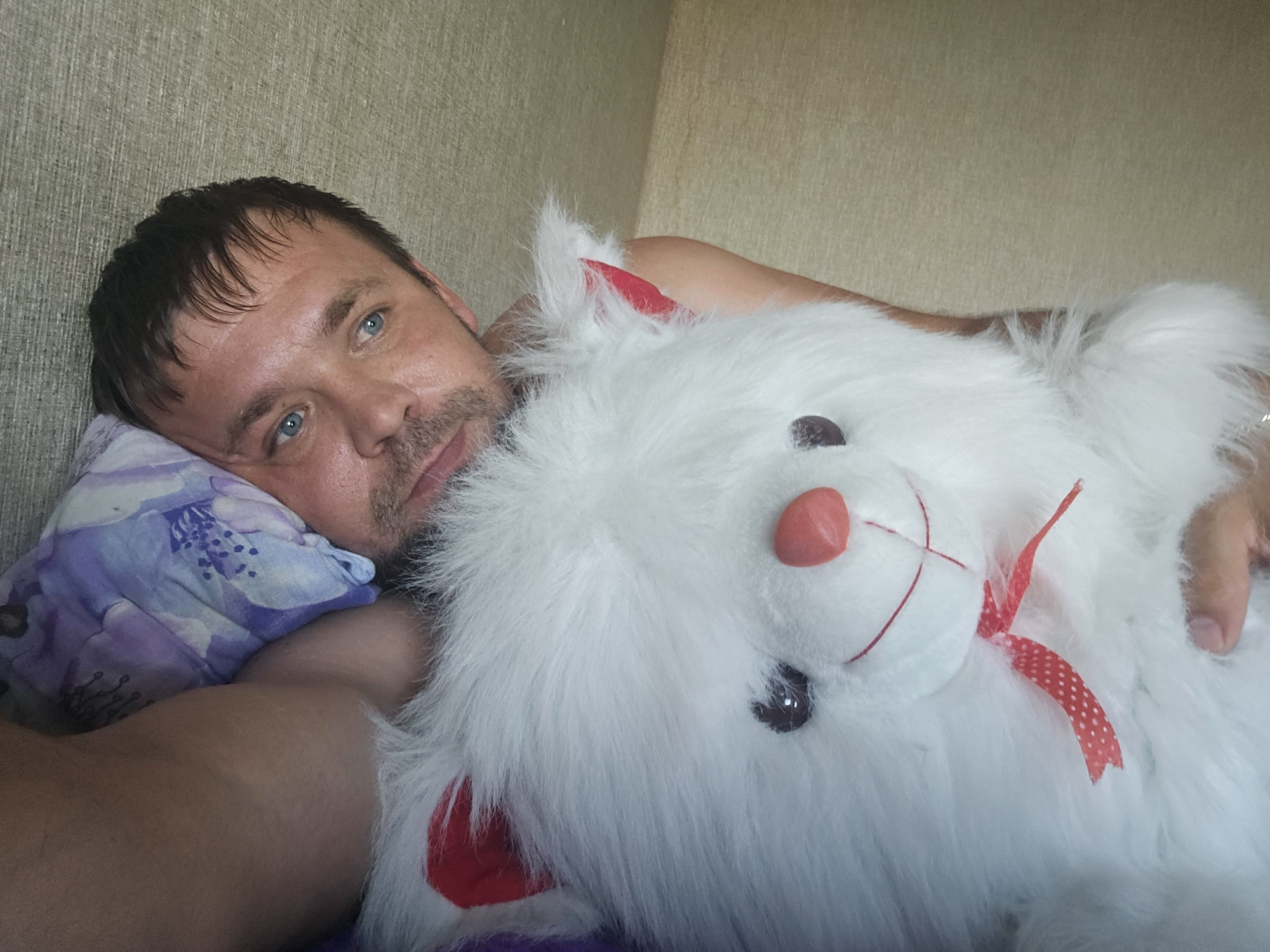 In the city of Taezhnoye, Russia, a close-up shot captures a man lying on his side, hugging a large white plush toy. The man, with short dark hair and blue eyes, rests his head on a pillow with a floral pattern. The plush toy, appearing to be a rabbit or similar creature, has a red nose, dark eyes, and a red ribbon tied around its neck. The background consists of a textured wall. The lighting suggests it is daytime.