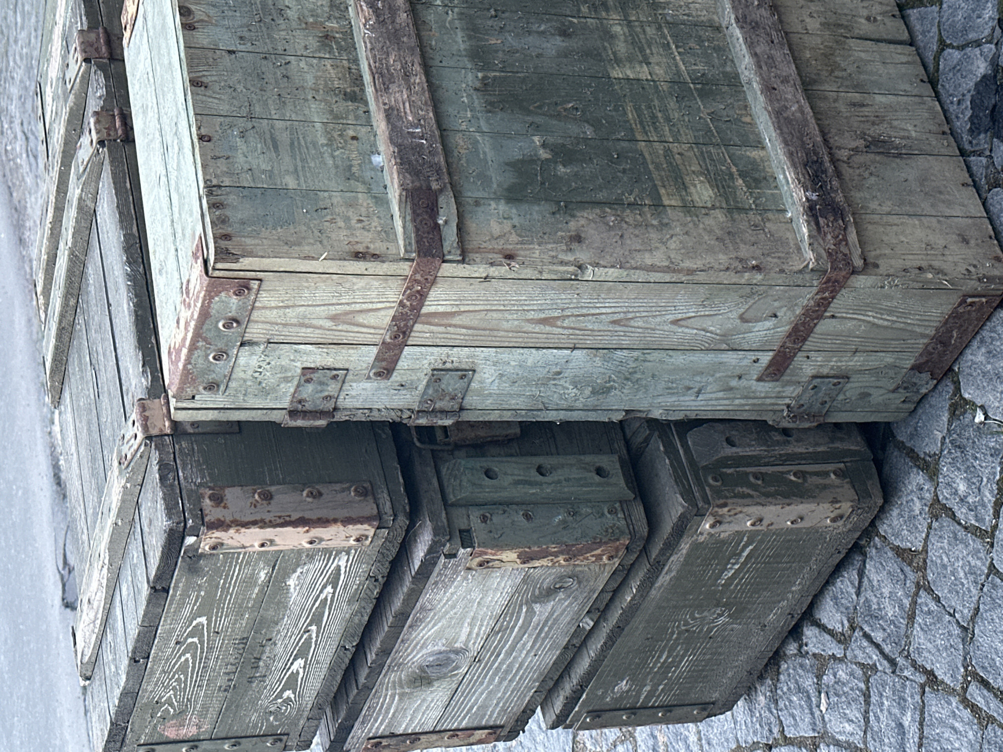 The image shows a stack of old, weathered wooden crates in Новоглаголево, Russia. The crates are likely military or industrial in origin, judging by their construction with metal strapping and corner reinforcements. The wood is aged and stained, and the metal hardware is rusty. They sit on a cobblestone or paved surface. The environment looks urban and perhaps slightly damp.