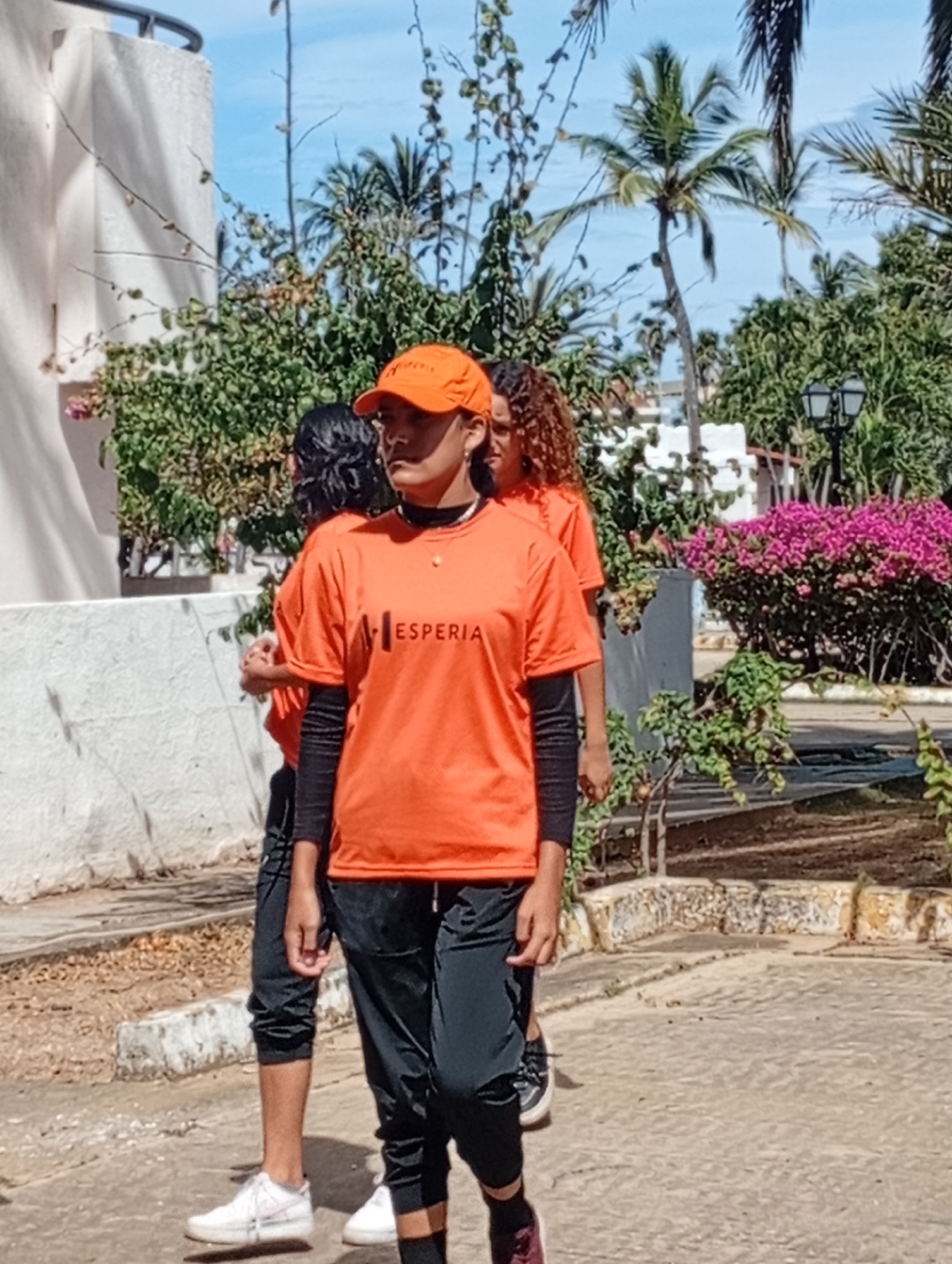 Three young women are walking in a park or plaza in Playa El Agua, Venezuela. They are wearing matching bright orange t-shirts with the word "ESPERIA" printed on the front, black long-sleeved shirts underneath, and dark pants. The woman in the front also wears an orange baseball cap. One woman has white sneakers, one has white sneakers, and one has dark sneakers. The background shows lush tropical vegetation, palm trees, flowering bushes, and white buildings. A street lamp is visible behind the women. The pavement is light-colored. The day is sunny.