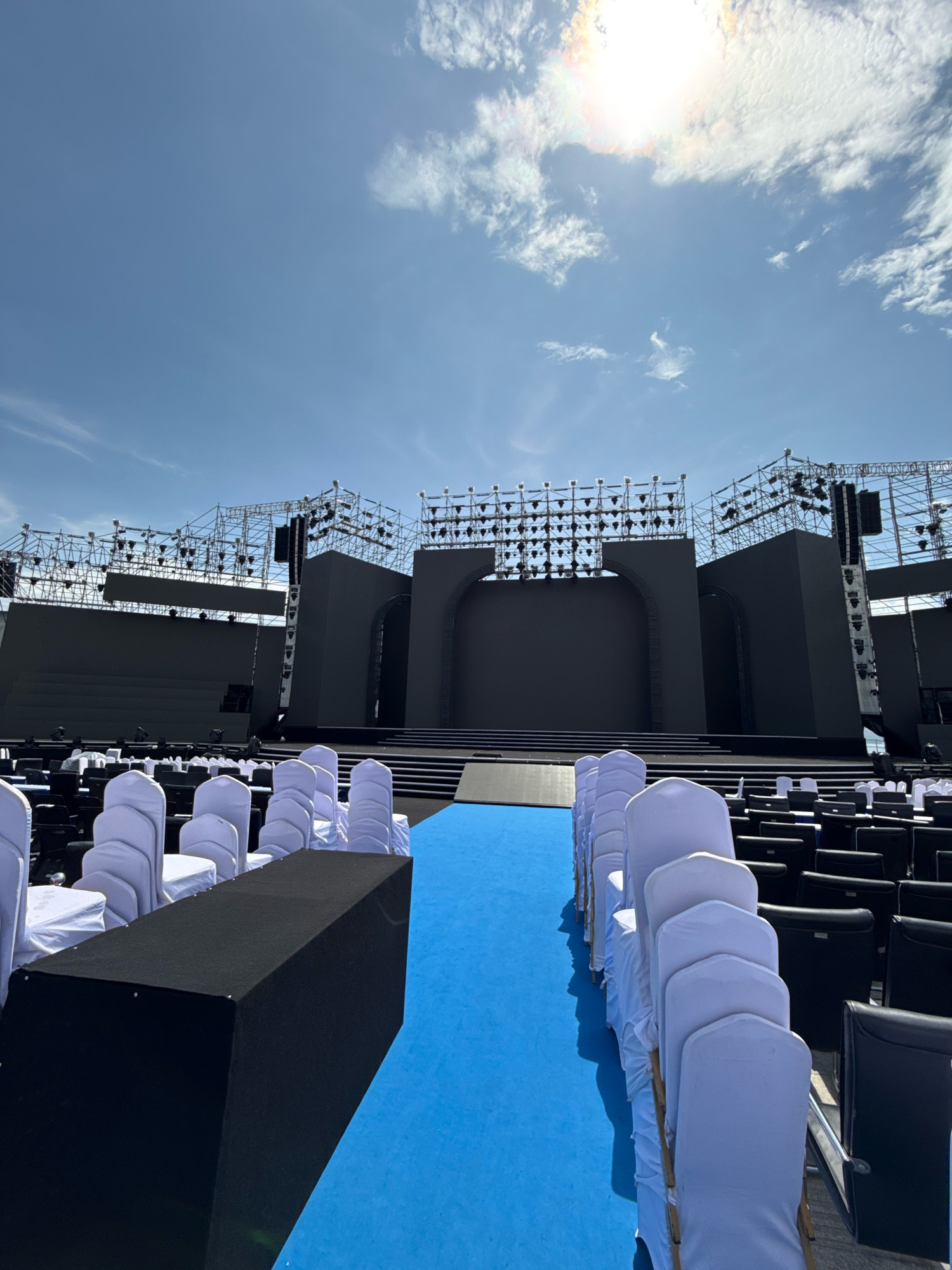 The image depicts an outdoor event setup, likely for a concert or performance, in Nha Trang, Vietnam. A large, dark stage structure dominates the background, with intricate scaffolding and lighting rigs overhead. In the foreground, rows of black and white chairs are arranged on a vibrant blue carpet. A black, rectangular platform is positioned to the left of the carpet. The sky above is clear blue with scattered white clouds, and the sun is visible, suggesting it is daytime. There are no people visible in the scene.