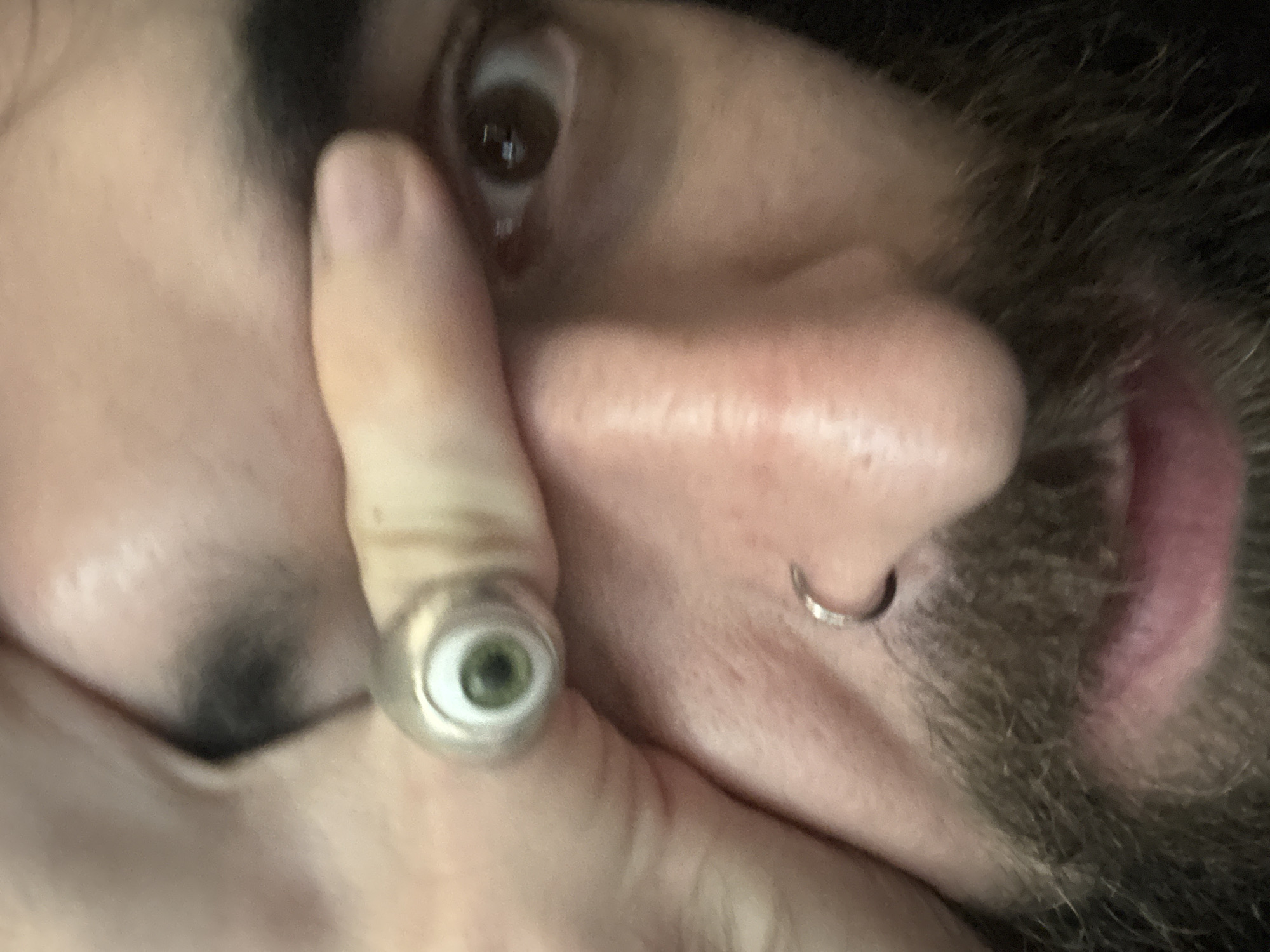 A close-up, low-angle shot shows a person's face from the nose up, with their hand covering their left eye and a ring on their index finger. The ring is silver with a green and black eyeball design. The person has dark hair and beard, and a nose ring. The background is dark and out of focus. The image does not contain any information or visual cues that would allow for the determination of its location in Phong Phú, Vietnam.