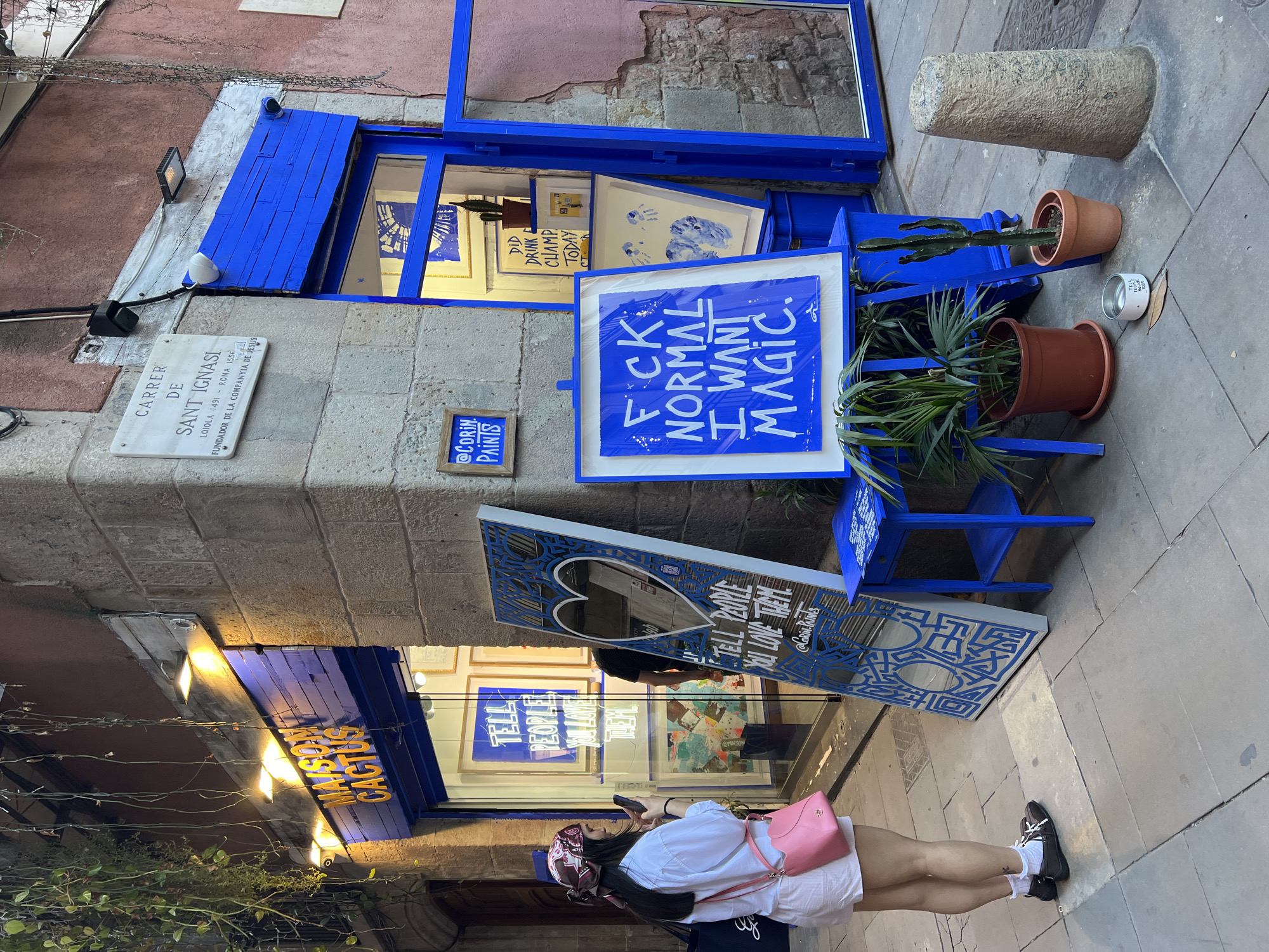 The image shows the exterior of a business named "Maison Cactus" located on Carrer de Sant Ignasi in Barcelona, Spain. A woman is standing in front of the store, looking at her phone, wearing a white shirt, white shorts, black shoes with white socks, a pink purse, and a bandana. The store has a blue-painted facade with multiple signs and artworks displayed outside. One sign says "FCK NORMAL I WANT MAGIC," another says "TELL PEOPLE YOU LOVE THEM." Potted plants, including a cactus, and a stone pillar are part of the storefront display. A plaque on the building identifies the street and includes historical information about Sant Ignasi.