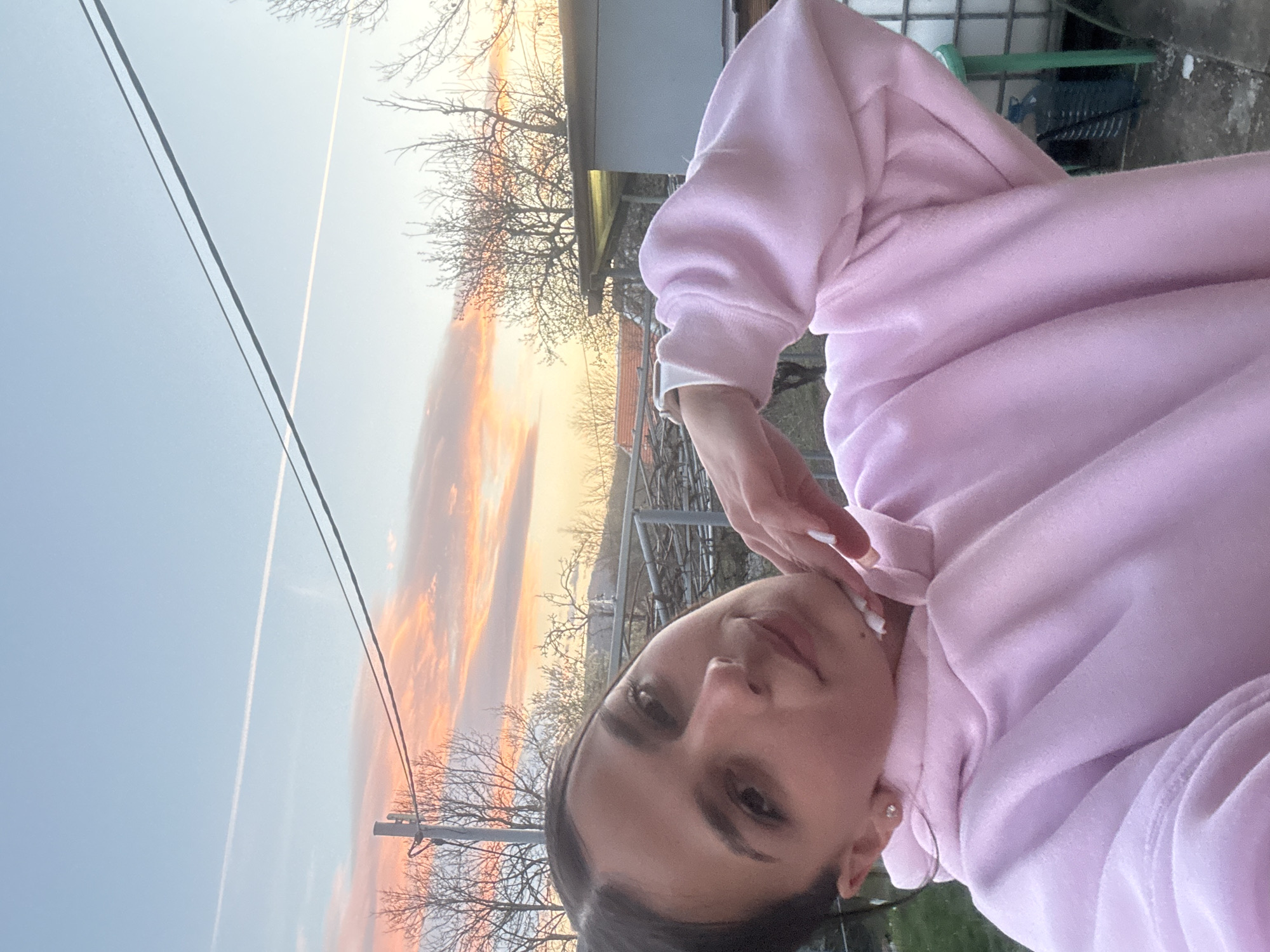 The image shows a young woman in a pink hoodie. She is facing the camera with her hand near her chin. In the background, there is a sunset sky with orange and yellow hues. Power lines and bare tree branches are visible. To the right of the sky, there is a building and green objects that appear to be part of the outdoor area of the house. It is hard to say the location is in Balinovtsi, Bulgaria based on the information given.