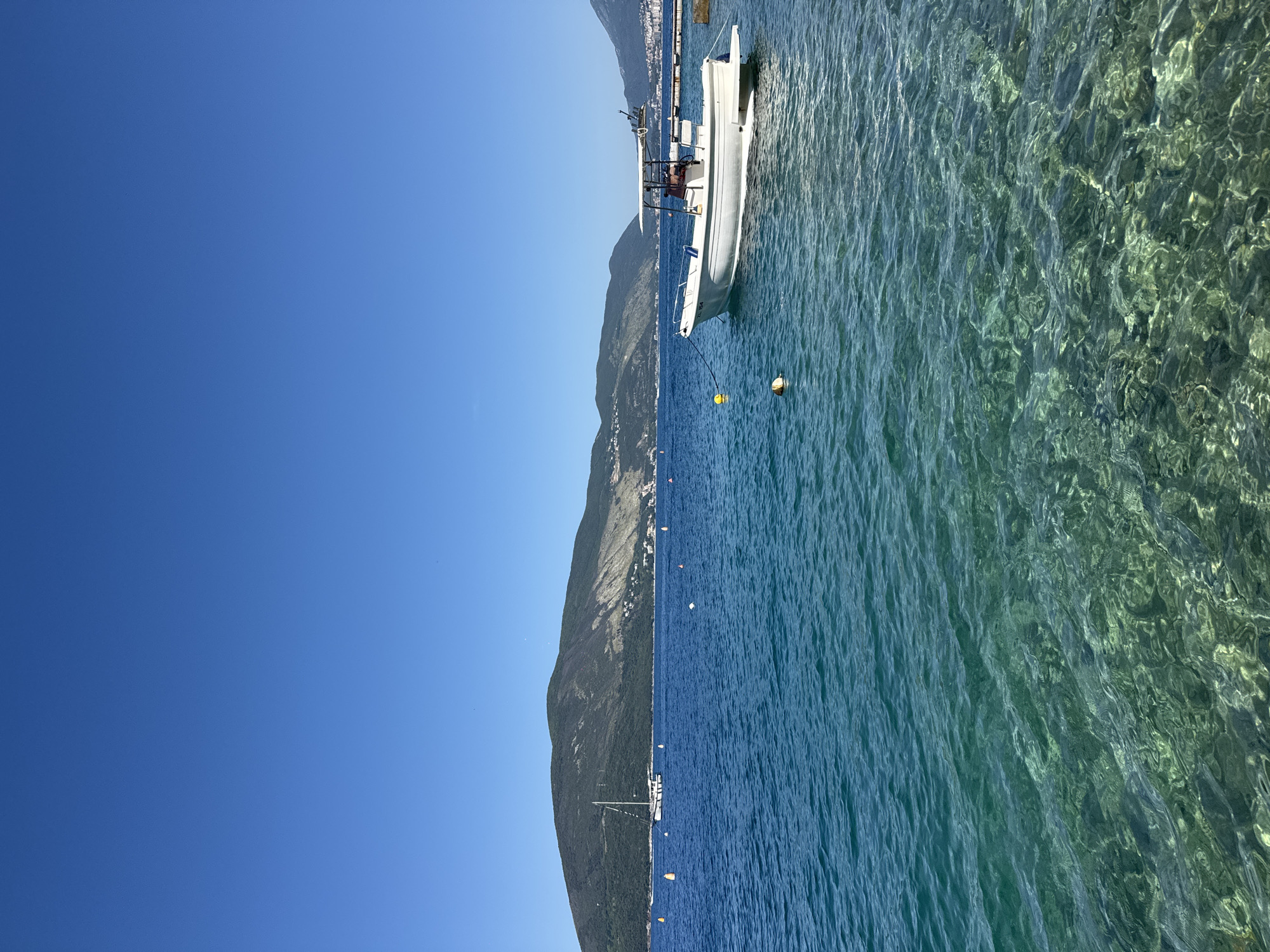 A vertical outdoor shot captures a vibrant coastal scene under a pristine, cloudless deep blue sky. The foreground features exceptionally clear, shallow turquoise water, revealing a rippled, rocky seabed with visible light patterns. The water gradually transitions to a deeper blue in the mid-ground. A white motorboat with a grey canopy, bearing the registration "ZC 154" on its hull, is moored to a yellow spherical buoy on the right side of the frame, adjacent to a small concrete pier. Further to the left, a white sailboat with a raised mast is anchored amidst several yellow and orange marker buoys. The background is dominated by a range of lush, dark green mountains, with clusters of white buildings visible on their lower slopes. Distant mountains extend into the hazy horizon on both sides. No individuals are distinctly visible.