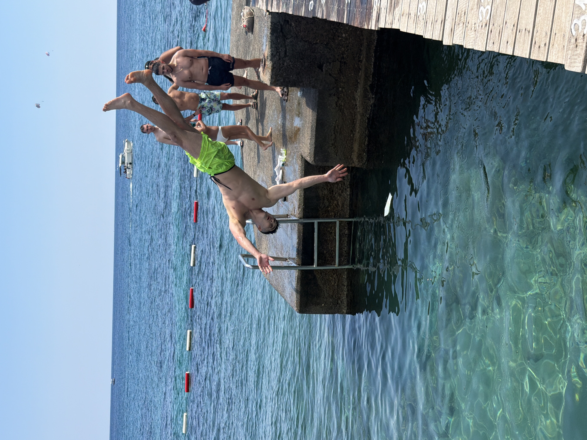 A young man in neon green swim trunks is captured mid-air in a diving pose, his body extended horizontally over the clear, turquoise water. He is diving off a concrete pier that has a metal ladder attached to its side, leading into the water. Behind him, on the pier, several other individuals are standing, observing the dive. The water is remarkably clear, allowing the seabed to be visible beneath the surface. In the background, the vast blue expanse of the Adriatic Sea stretches towards the horizon, with a few boats dotting the water and two parasails visible in the sky. The scene is bathed in bright sunlight, suggesting a warm, summery day. The concrete pier appears to be a popular spot for diving, likely in Budva, Montenegro, given the characteristic clear waters and the style of the pier.