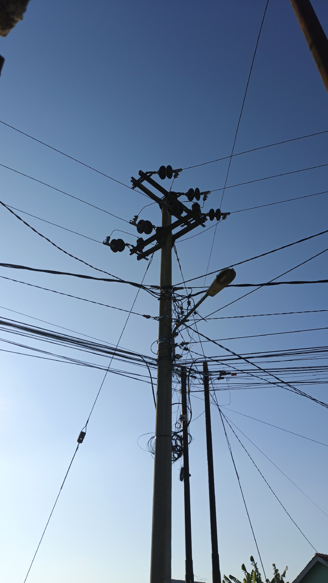 A low-angle, medium shot captures a utility pole against a clear blue sky in Santing, Indonesia. The concrete pole, situated in the center of the frame, is heavily laden with a dense network of tangled electrical wires. Attached to the pole are multiple insulators and what appear to be transformers. A street lamp is also affixed to the pole, extending outwards. To the right of the main pole, two thinner, darker poles are visible, also supporting overhead wires. In the bottom right corner, a portion of a house roof and some green foliage are visible. The overall impression is one of a cluttered, yet functional, urban infrastructure against a serene sky.