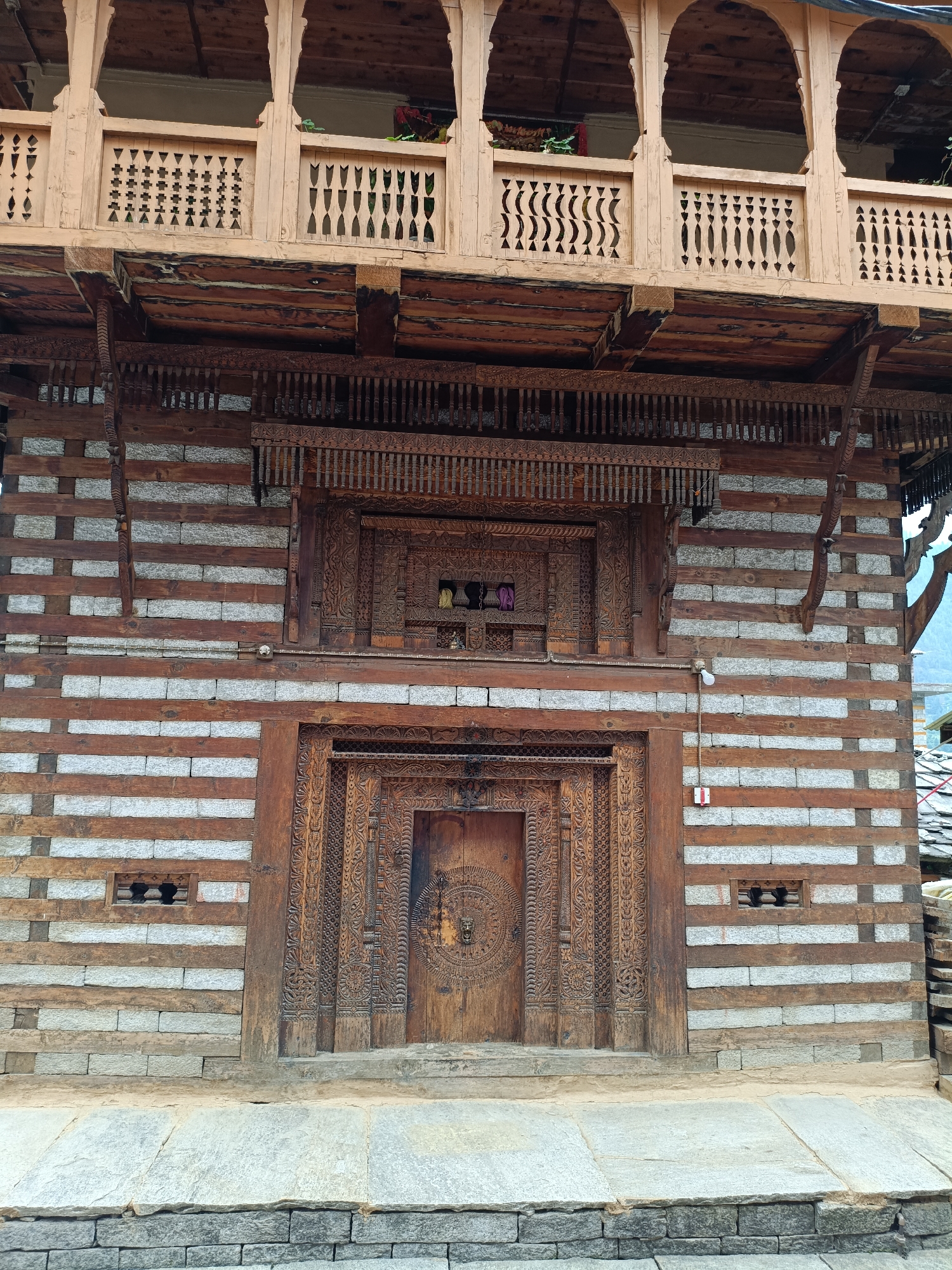 The image shows the exterior of a traditional wooden and stone building in Vashisht, India. The architecture features intricate wood carvings on the doors and balconies, with horizontal bands of wood and stone making up the walls. A large, ornately carved wooden door is centrally located on the lower level. Above the door is a smaller, carved wooden structure with some items visible inside. The upper level has a wooden balcony with arched openings and carved railings. The building appears old and weathered, with evidence of traditional craftsmanship. The surrounding environment is not fully visible, but some glimpses of a tiled roof and mountains in the distance can be seen. There are no people visible in the image.