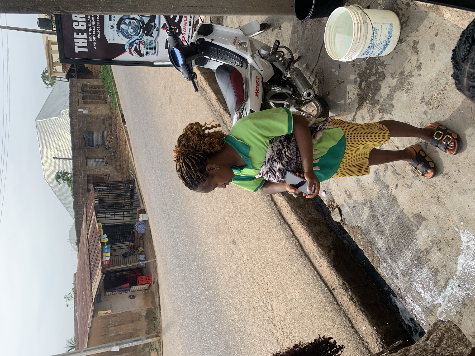 A woman with braided hair, wearing a light green polo shirt and a yellow patterned skirt, stands on a concrete sidewalk holding a black smartphone. She has a patterned cloth wrapped around her shoulders. Next to her, a white motorcycle with red accents is parked. Behind the motorcycle is a tall, dark sign advertising "THE GR..." services, including "BOUTIQUE, LAUNDRY AND DRY CLEANING S..." and a phone number, along with an image of a washing machine. To the left of the woman is a deep drainage ditch with some water and foam. Further left, a small storefront with a rusted tin roof is visible, displaying red crates and various goods behind a metal gate. In the background, buildings constructed with brick and corrugated metal roofing are present, along with a large, white, angular structure, possibly a covered market or greenhouse. The scene appears to be on a street in Jos, Nigeria, characterized by its urban infrastructure, commercial activities, and the presence of local businesses.