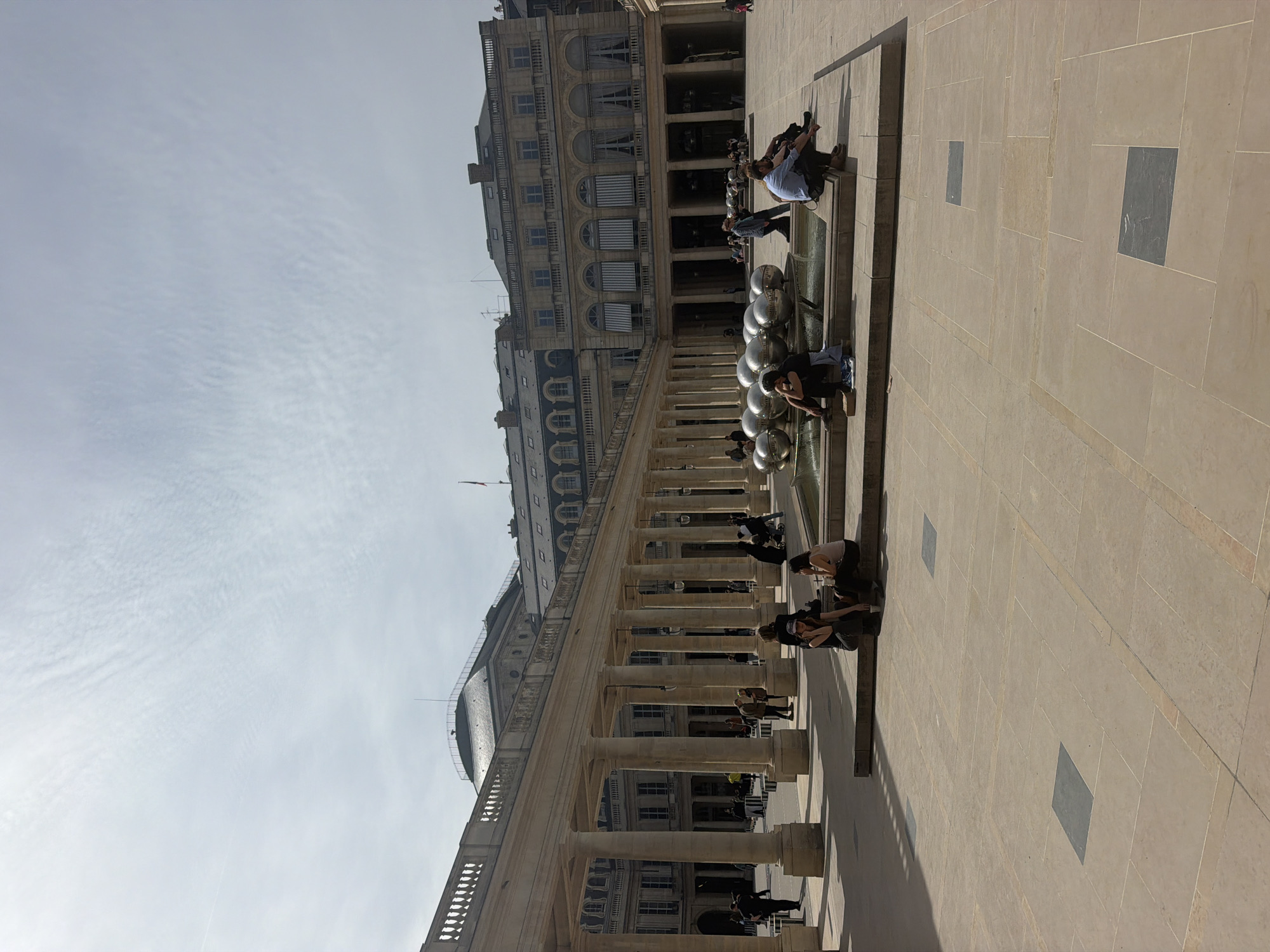 The photograph captures a view of the Palais Royal in Paris, France. The architecture showcases the iconic colonnades and buildings surrounding the courtyard. A unique modern fountain with silver spheres is present in the foreground. Several people are sitting and interacting with the fountain. The pavement is visible with square inlays. The sky is partly cloudy.