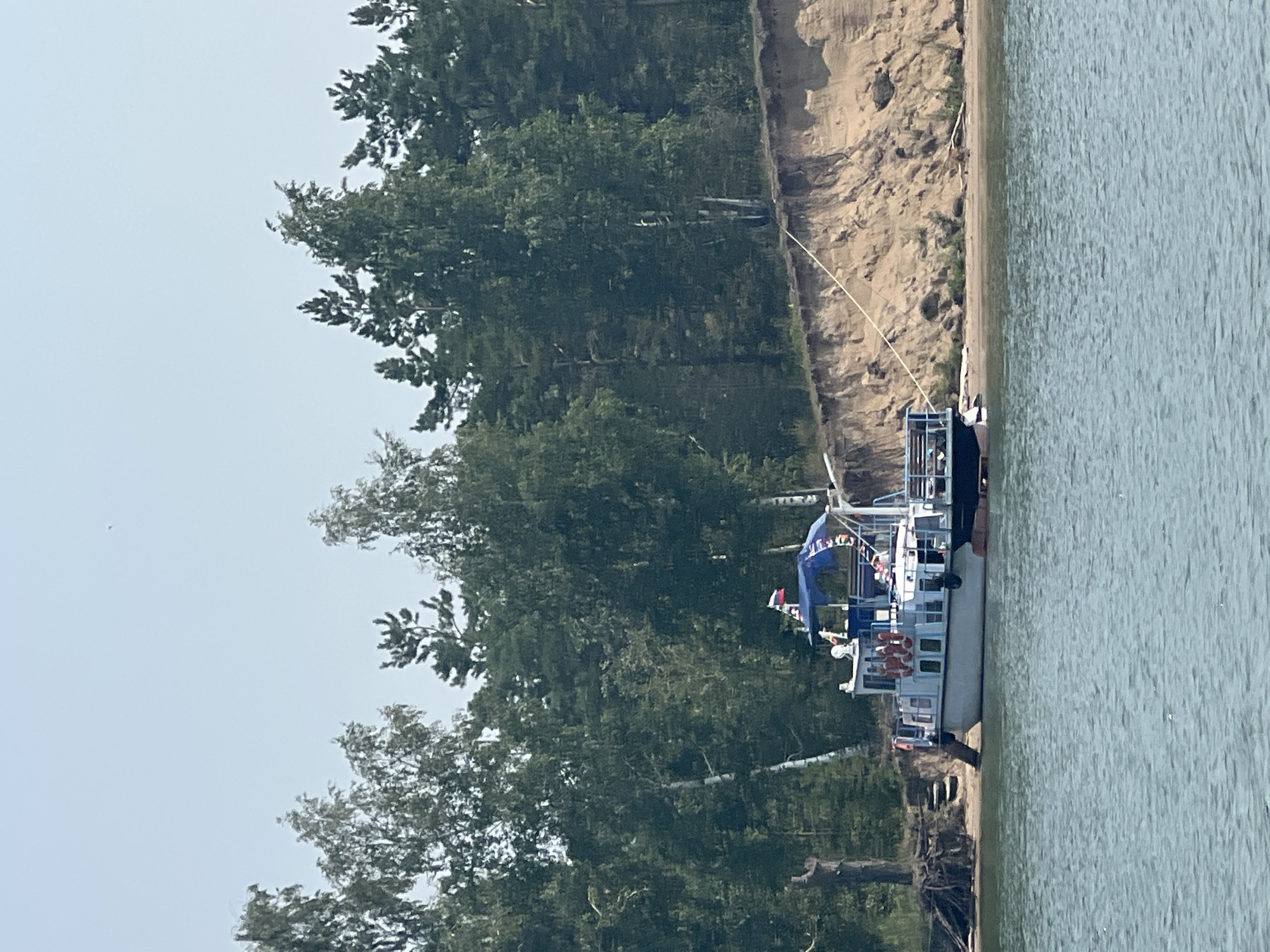 A wide shot captures a serene waterfront scene in Borovoe, Russia, dominated by a calm, rippling body of water in the foreground. The water transitions to a sandy embankment that rises to a dense forest of tall, green trees. On the sandy shore, a white and blue passenger boat is docked, its roof adorned with a Russian flag. The boat appears to be unoccupied. A dark, smaller boat is moored to the side of the larger vessel. The embankment shows signs of erosion, with visible layers of soil and sand, and a thin rope runs from the boat to the top of the embankment. The sky is a pale, hazy blue, indicating fair weather. The overall impression is one of a peaceful, natural setting.