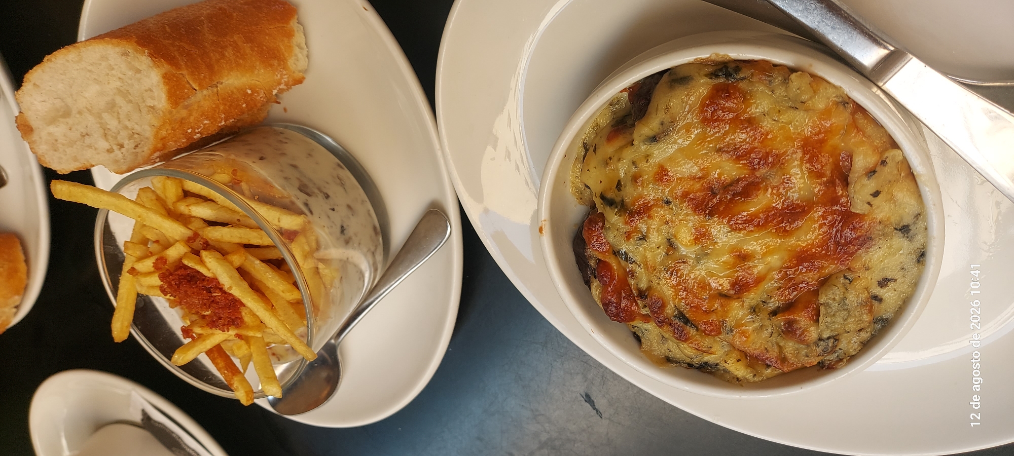 The image depicts a close-up of two plates of food, likely served in a restaurant in Pamplona, Spain.
The upper plate features a clear glass dish filled with french fries, topped with a sprinkle of crumbled bacon or ham. Beneath the fries, a creamy sauce with visible mushroom pieces can be seen. To the right of the dish is a piece of crusty bread, partially eaten. A spoon rests on the plate, positioned to be used with the dish.
The lower plate holds a white ceramic ramekin filled with a baked dish, characterized by a generous topping of melted, golden-brown cheese. The cheese appears bubbly and slightly browned in places, indicating it has been broiled or baked at a high temperature. Beneath the cheese, darker elements are visible, possibly spinach or other greens mixed with a creamy base. A silver utensil, likely a knife or a large fork, is placed to the right of this dish.
The background suggests a dark tabletop, with the white plates standing out. A timestamp in the lower left corner reads "12 de agosto de 2026 10:41," indicating the date and time the photograph was taken. The overall presentation suggests a casual dining experience, possibly tapas or appetizers. There are no visible people in the frame.