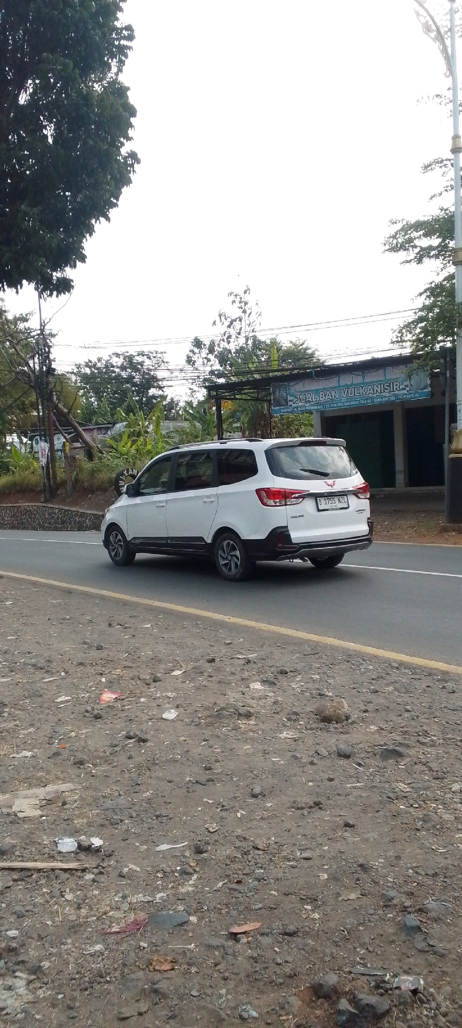 A white four-door SUV is traveling on a paved road in Cilaku, Indonesia. The vehicle has a license plate that reads "B 1755 NZL." The road is lined with a yellow painted line and a rough, rocky shoulder. In the background, there is a building with a banner that says "JUAL BAN VULKANISIR." The area appears to be a commercial or industrial zone, with some trees and vegetation visible. The sky is overcast.