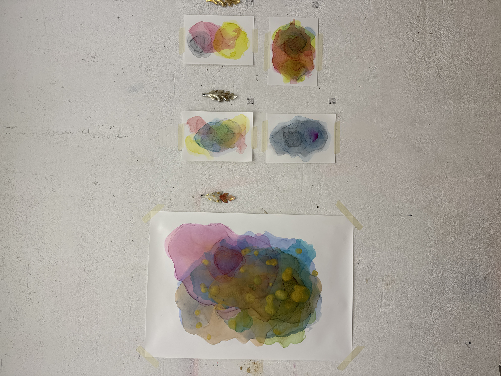 The image shows a section of a white, textured wall, likely indoors. Affixed to the wall with tape are several abstract watercolor paintings on white paper. These paintings feature overlapping blotches of color, including pink, yellow, green, blue, and gray. Above some of the paintings are decorative metal leaf-shaped ornaments. The image does not contain sufficient information to determine a specific location in Moscow, Russia.