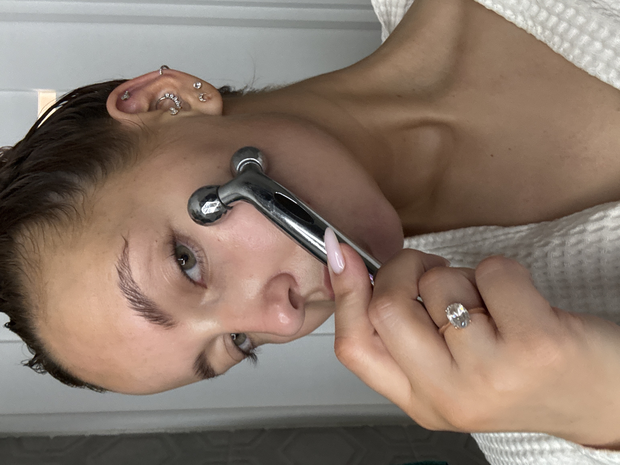 A woman is using a facial massager on her cheek. She is wearing a white robe and has several ear piercings. She is also wearing a ring on her finger. The location cannot be determined as Chaikovsky, Russia. The environment appears to be an indoor setting.