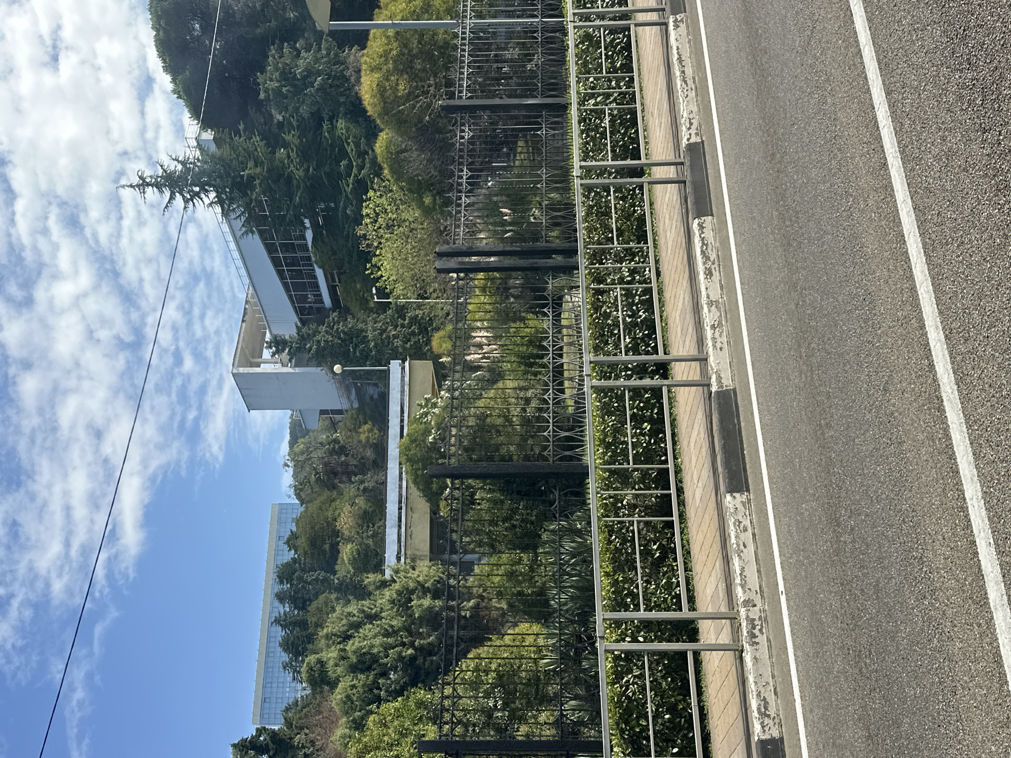 The image depicts a view of the Zhemchuzhina Hotel complex in Sochi, Russia, on a clear day with scattered clouds. In the foreground, an asphalt road with white lane markings is visible, bordered by a sidewalk with a metal railing and a low hedge. Behind the hedge, a decorative wrought iron fence encloses a lush garden area with various trees and shrubs. Beyond the fence, several buildings are situated on a sloping hill. A modern, multi-story building with a glass facade is visible on the left, surrounded by evergreen trees. To the right of this, a white, angular structure with a sloping roof and large windows appears to be part of a recreational facility or an extension of the hotel. The hillside is densely covered with mature trees, creating a verdant backdrop against the blue sky. The overall scene conveys a sense of an established resort area with a blend of modern architecture and natural landscape. There are no people visible in the image.