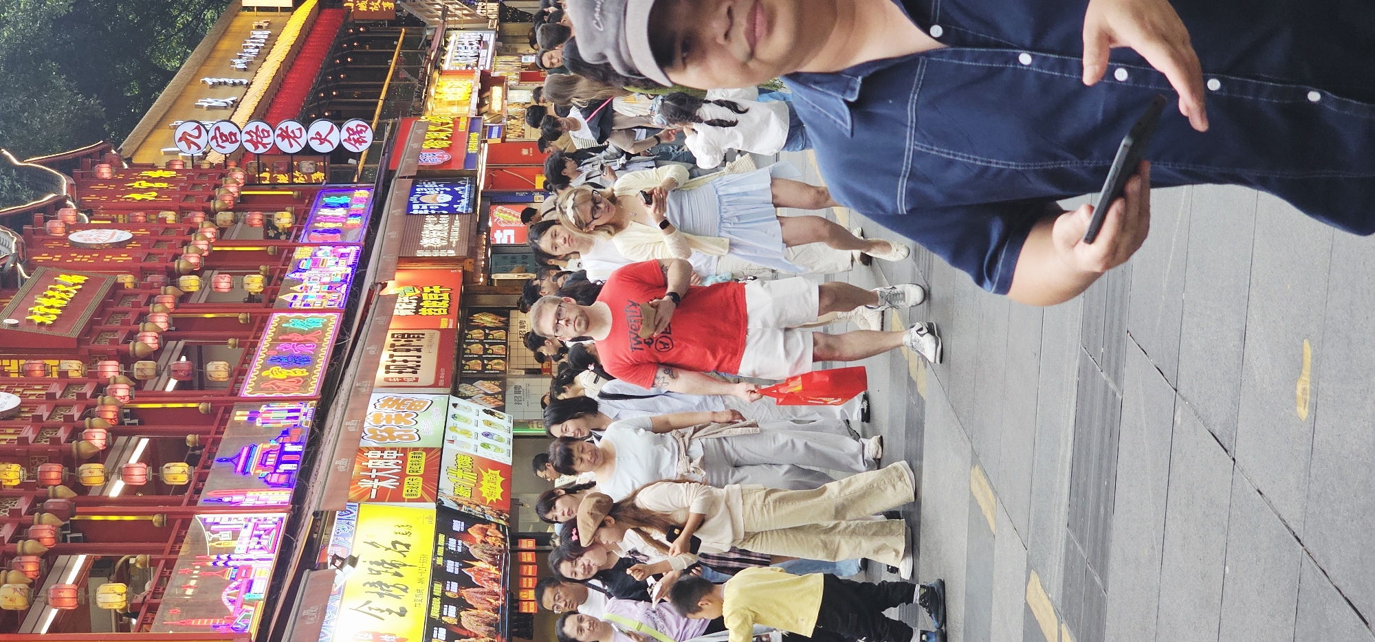 The image depicts a busy street scene in Yuzhong District, China. A crowd of people stands on a paved sidewalk lined with vibrant storefronts. The storefronts are adorned with numerous brightly lit signs, many written in Chinese characters, creating a visually stimulating environment. The people are dressed in casual clothing, indicative of everyday life or tourism. One man in the foreground wears a blue shirt and a cap, partially obscuring the view. Another man stands out in a red t-shirt and white shorts. Some people are looking at their phones. The environment appears to be bustling with activity, indicative of a lively urban atmosphere in a commercial area of Yuzhong District. The presence of lanterns suggests a cultural or festive element.