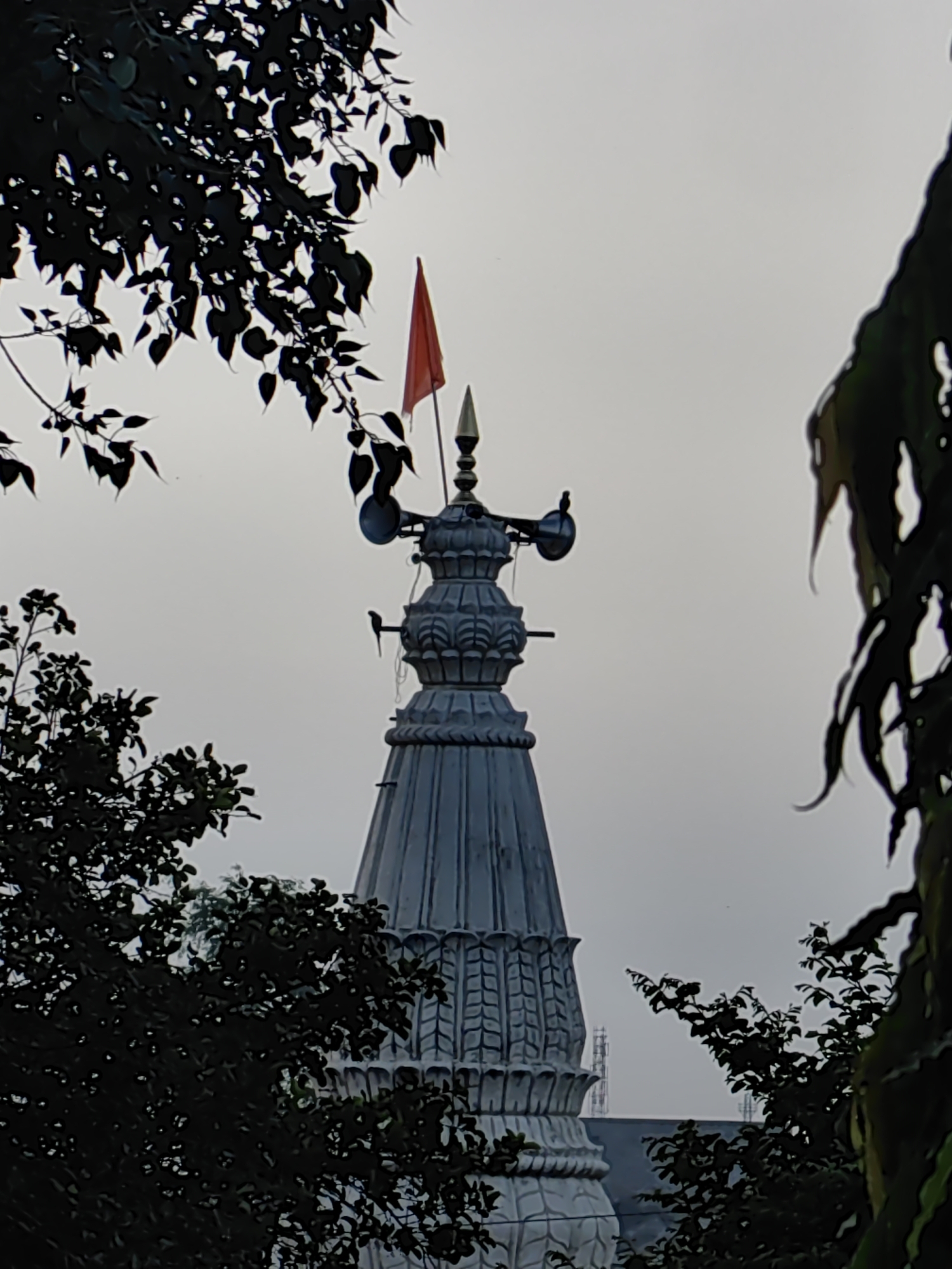 The image shows the spire of a temple in Mhaiswadi, India. The spire is made of grey stone and is decorated with intricate carvings. A red flag is flying from the top of the spire. There are trees in the foreground and background, and the sky is overcast. The image appears to have been taken from a distance, possibly from across a street or a body of water. The overall mood of the image is serene and peaceful.