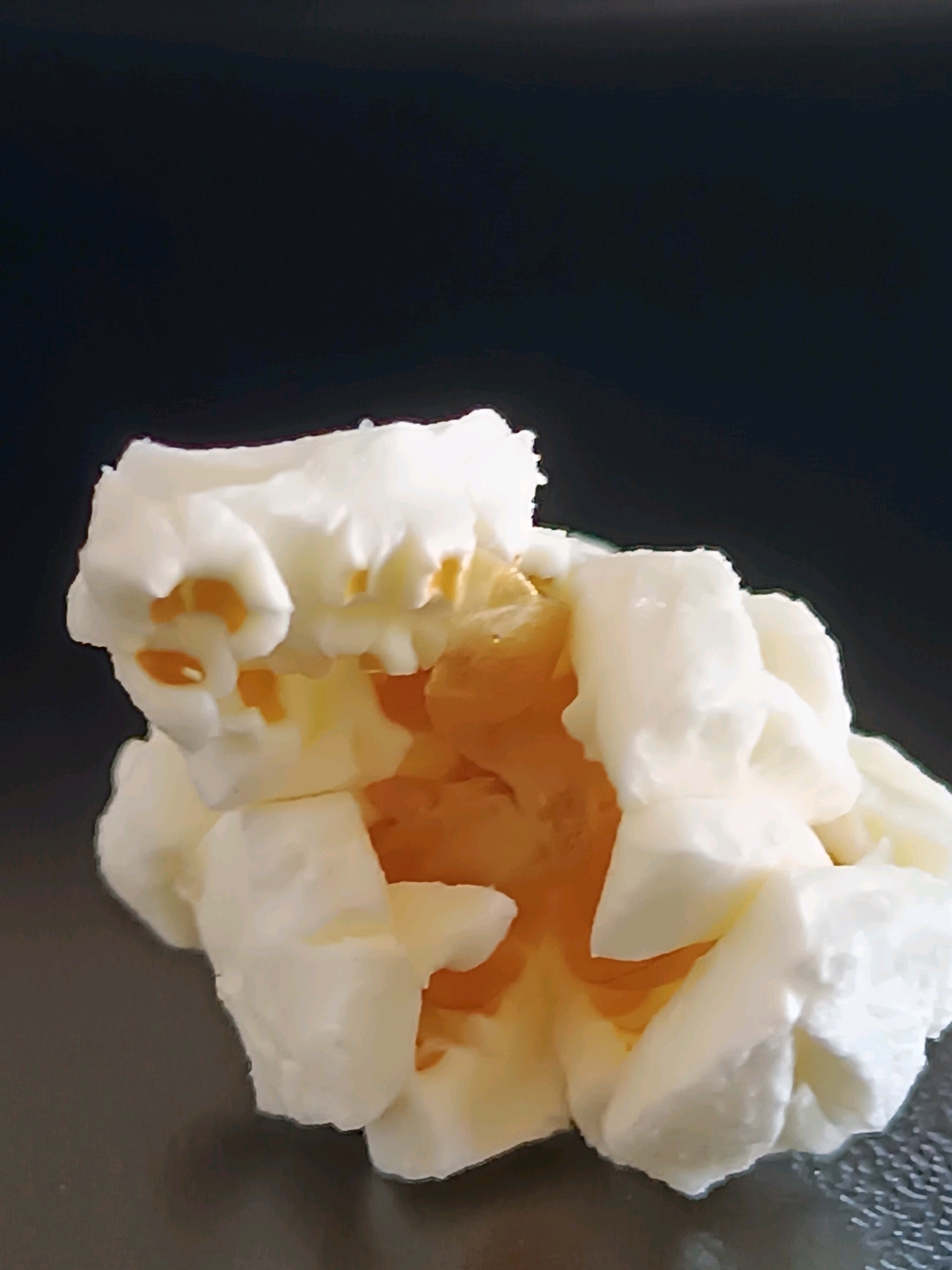 A close-up, low-angle shot shows a clump of white popcorn against a dark background. The popcorn is fluffy and irregular in shape, with some pieces appearing to be partially popped or broken. The inner parts of the popcorn are visible, revealing a translucent, golden-yellow hue. The lighting highlights the texture and curves of the popcorn. The image's composition is tight, focusing entirely on the popcorn itself, with no surrounding environment visible. There is no indication of the city of Viadana, Italy, or any other geographical location. The scene is static and there are no people or actions depicted.