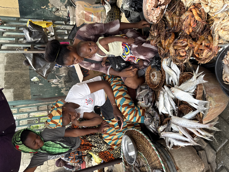 This image depicts a vibrant outdoor market scene in Lekki, Nigeria, focused on seafood sales.
**Location:** Lekki, Nigeria. The specific street or market name is not identifiable within the image, but it is an urban outdoor setting.
**Scene Overview:** The foreground is dominated by an abundant display of fresh seafood, primarily various species of fish and prawns, arranged in numerous woven baskets and metal containers on the ground. Several individuals, predominantly women, are present, engaged in market activities.
**People:**
* **Foreground Left:** A young woman stands, wearing a grey t-shirt and a patterned green headscarf with a distinct logo (resembling Gucci). Her skirt features a vibrant orange and black geometric pattern. She smiles downwards, her left hand resting on an unseen object under a purple market umbrella.
* **Foreground Center:** A woman sits, dressed in a white t-shirt with a "Corinthians" logo and patterned trousers featuring brown, blue, and white geometric shapes. Her head is covered with a matching patterned head wrap. She looks down at a pink smartphone held in her hands.
* **Foreground Right:** Another woman sits, wearing a brown and white patterned dress, with her dark hair styled upwards in a bun. She holds a young child on her lap. The child, dressed in a light yellow sleeveless top and patterned shorts (red, blue, yellow), looks towards the viewer. The woman has a black shoulder bag resting on her lap.
* **Background Left:** Partially visible, another woman sits on the ground amidst goods, wearing patterned clothing.
* **Background Right:** A blurry figure is partially visible.
**Objects and Subjects:**
* **Seafood:** Numerous types of fish, including silver-scaled fish, darker grey fish, and various sizes of prawns/shrimp, are neatly arranged in approximately ten round, woven baskets and a large rectangular metal pan. Some fish are displayed directly on a surface.
* **Market Equipment:** A large, round woven basket contains a metal pan. A dark purple market umbrella provides shade from above.
* **Background Items:** A "POP" branded cardboard box is visible behind the sitting woman in the center. A white woven bag, possibly containing "RIA CEMENT," is behind the woman holding the child. A black plastic bag is near the child.
**Environment:** The ground consists of grey interlocking paving stones. The background features a distressed wall, painted green with visible yellow patches and signs of wear. A green metal gate or fence structure is also visible against the wall. The lighting suggests daytime.