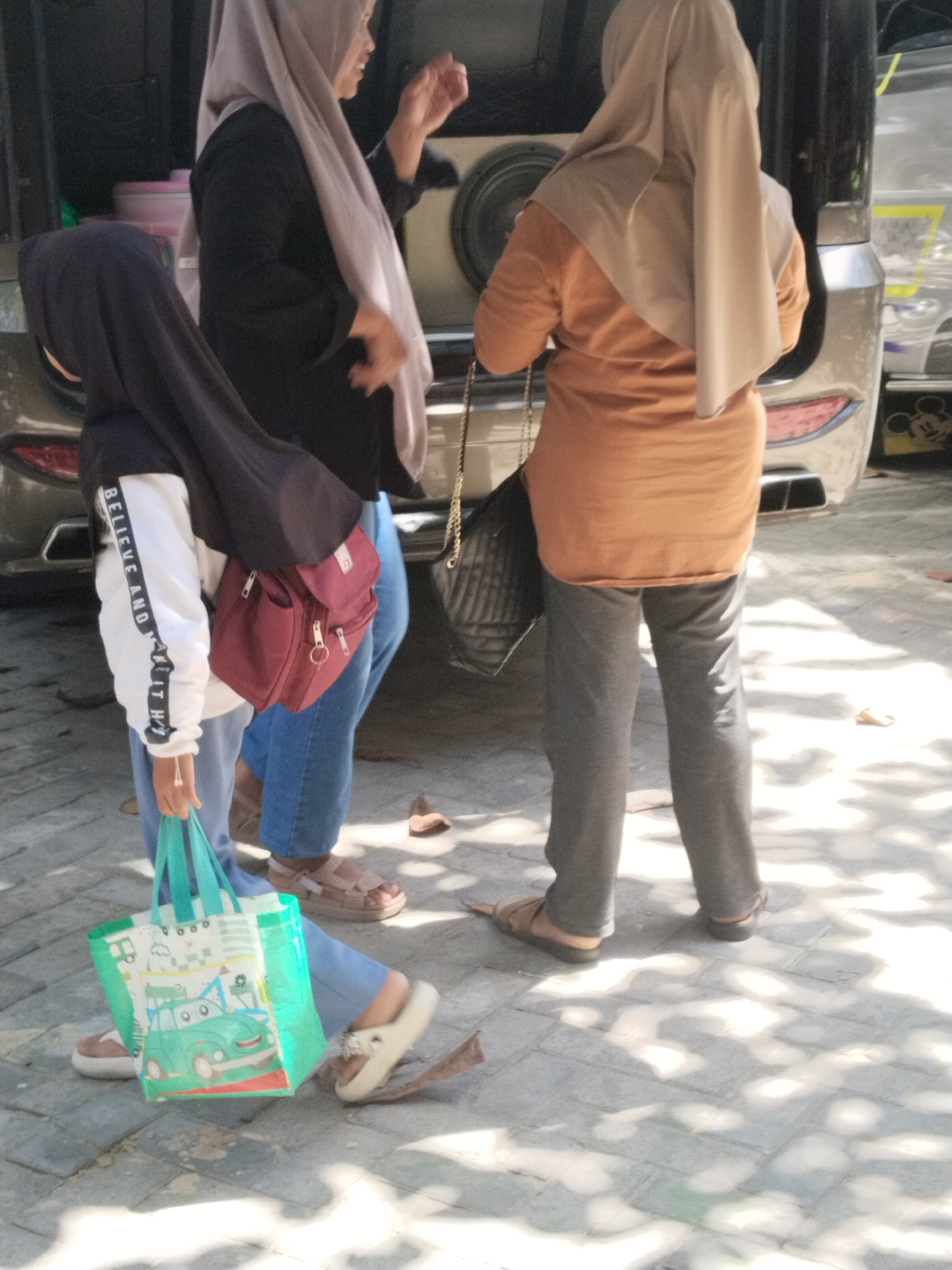 In Prigi, Indonesia, three individuals are standing on a paved surface, possibly a sidewalk or parking lot, near a vehicle. The person in the foreground, on the left, is wearing a black hijab and a white and black long-sleeved shirt with the words "BELIEVE AND MAKE IT HAPPEN" printed vertically on the sleeve. This individual is also holding a green reusable shopping bag with a cartoon image of a car. They are wearing light blue pants and white sandals.
To their right, a person in a black hijab and a light grey scarf is wearing a black long-sleeved shirt and blue jeans. They have a maroon crossbody bag.
On the far right, a person wearing a light brown hijab and an orange long-sleeved shirt is wearing grey pants and brown sandals. They are carrying a black quilted bag with a chain strap. The background shows parts of a vehicle, including its bumper and a large speaker. The ground is illuminated by dappled sunlight, creating shadows from overhead foliage.