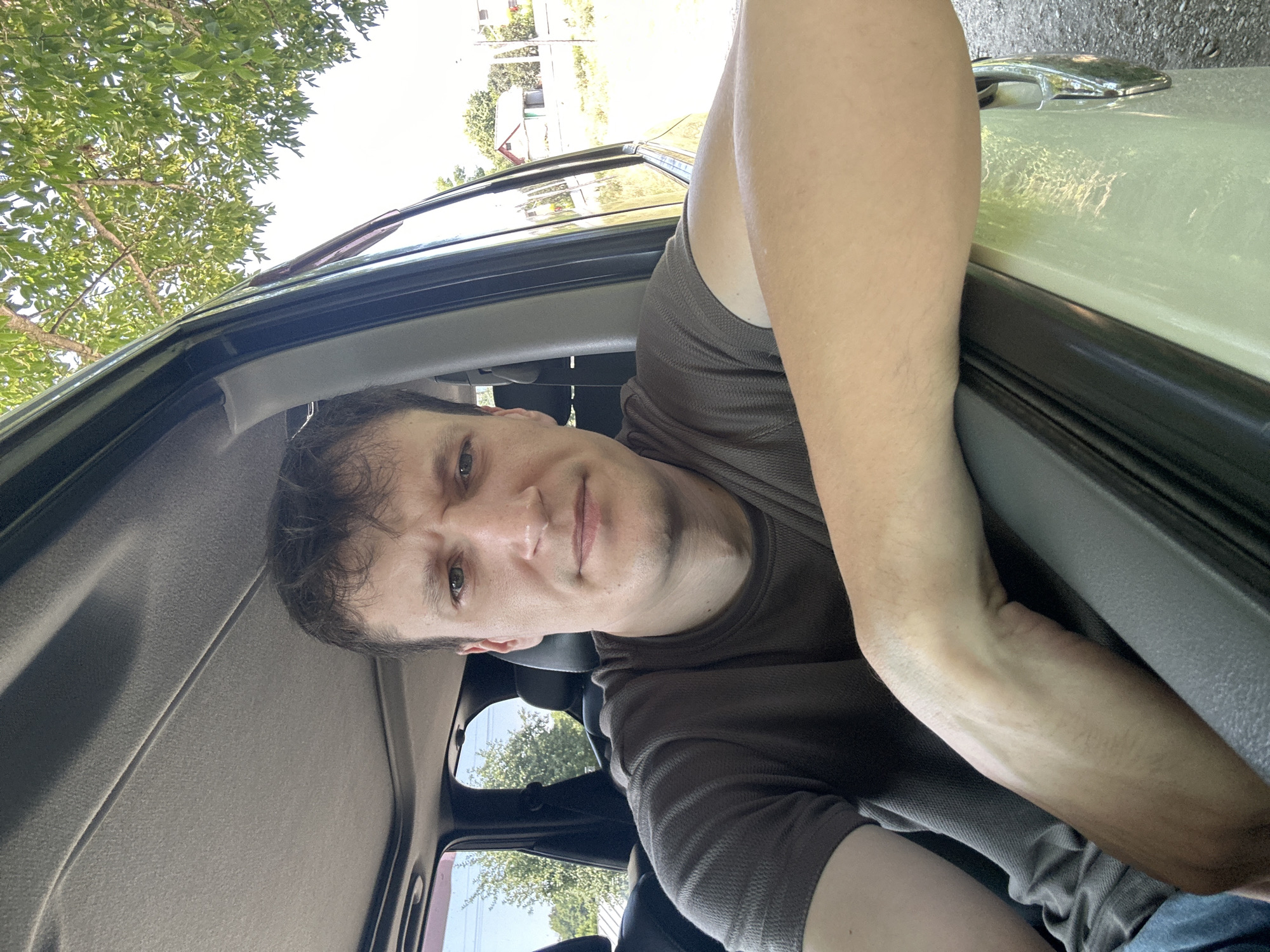 The image shows a man sitting in the driver's seat of a car, looking at the camera. He is wearing a brown t-shirt and has short, brown hair. His left arm is resting on the open car door, and his right arm is not visible. The car appears to be parked outdoors, with trees and a house visible in the background. The lighting suggests it is daytime. The location of Makiv, Ukraine, is not depicted in the image and cannot be inferred from the visual information provided.