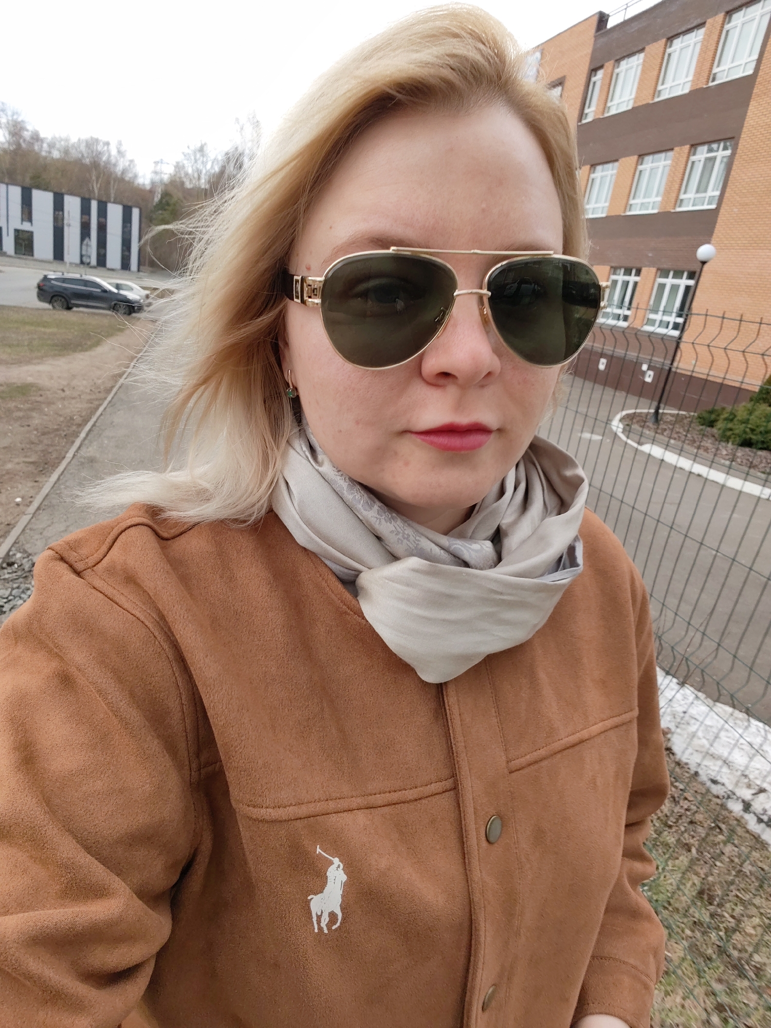 A woman with blonde hair and sunglasses is taking a selfie outdoors. She is wearing a brown jacket with a white logo of a person on horseback, and a light-colored scarf.
Behind her, there is a building with a brick facade and several windows. To the left, there is a building with black and white vertical stripes, and parked cars are visible in front of it. A gray fence is present in the right foreground, with patches of snow on the ground. The location appears to be Izhevsk, Russia, based on architectural styles and surroundings.