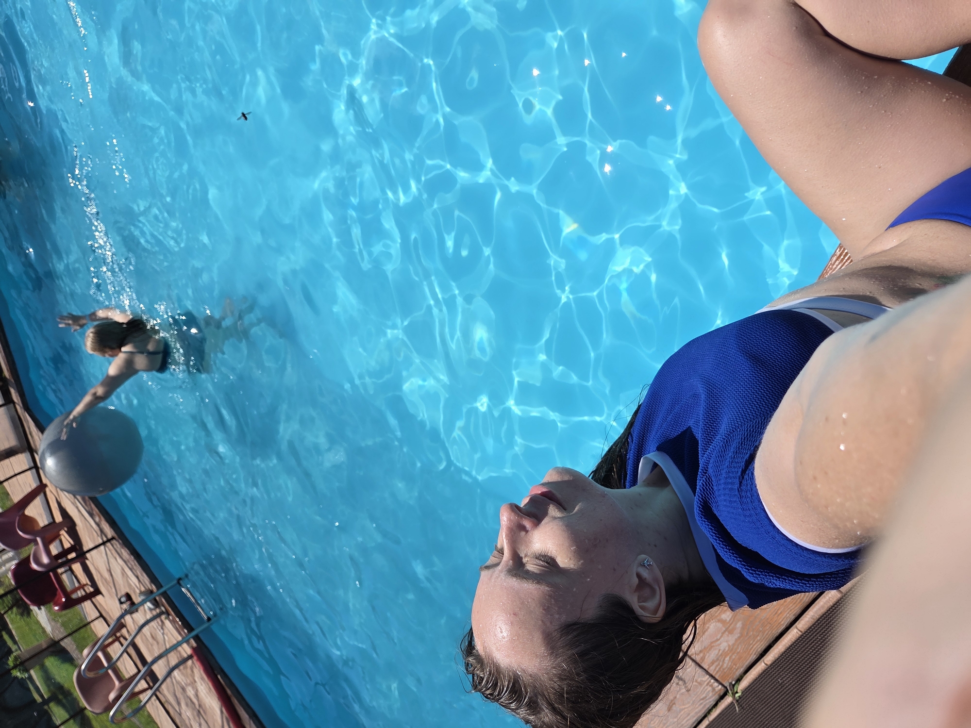 The image shows two young women in a swimming pool on a sunny day. In the foreground, a woman with her eyes closed and head tilted back is lying on the edge of the pool, partially in the water. She is wearing a blue bikini top. In the background, another woman is swimming in the pool, holding a large gray ball. The pool is surrounded by a wooden deck and lounge chairs. The location is not specified as Birobidzhan, Russia; this information cannot be determined from the image.