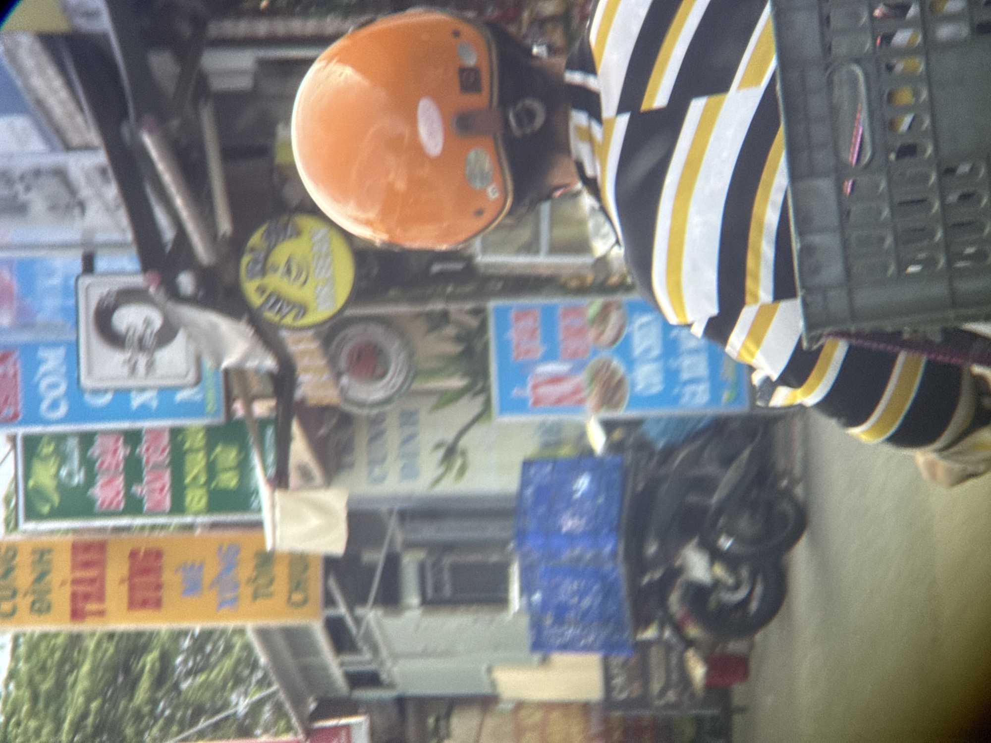 A person wearing a striped shirt and an orange helmet is riding a motorcycle in An Cựu Ward, Vietnam. The person is holding a black basket. In the background, there are several signs, including a yellow sign with Vietnamese text, a green sign with Vietnamese text, and a blue sign with Vietnamese text. There are also two blue crates on the back of another motorcycle. The scene appears to be a street in a city, with buildings and trees visible.
