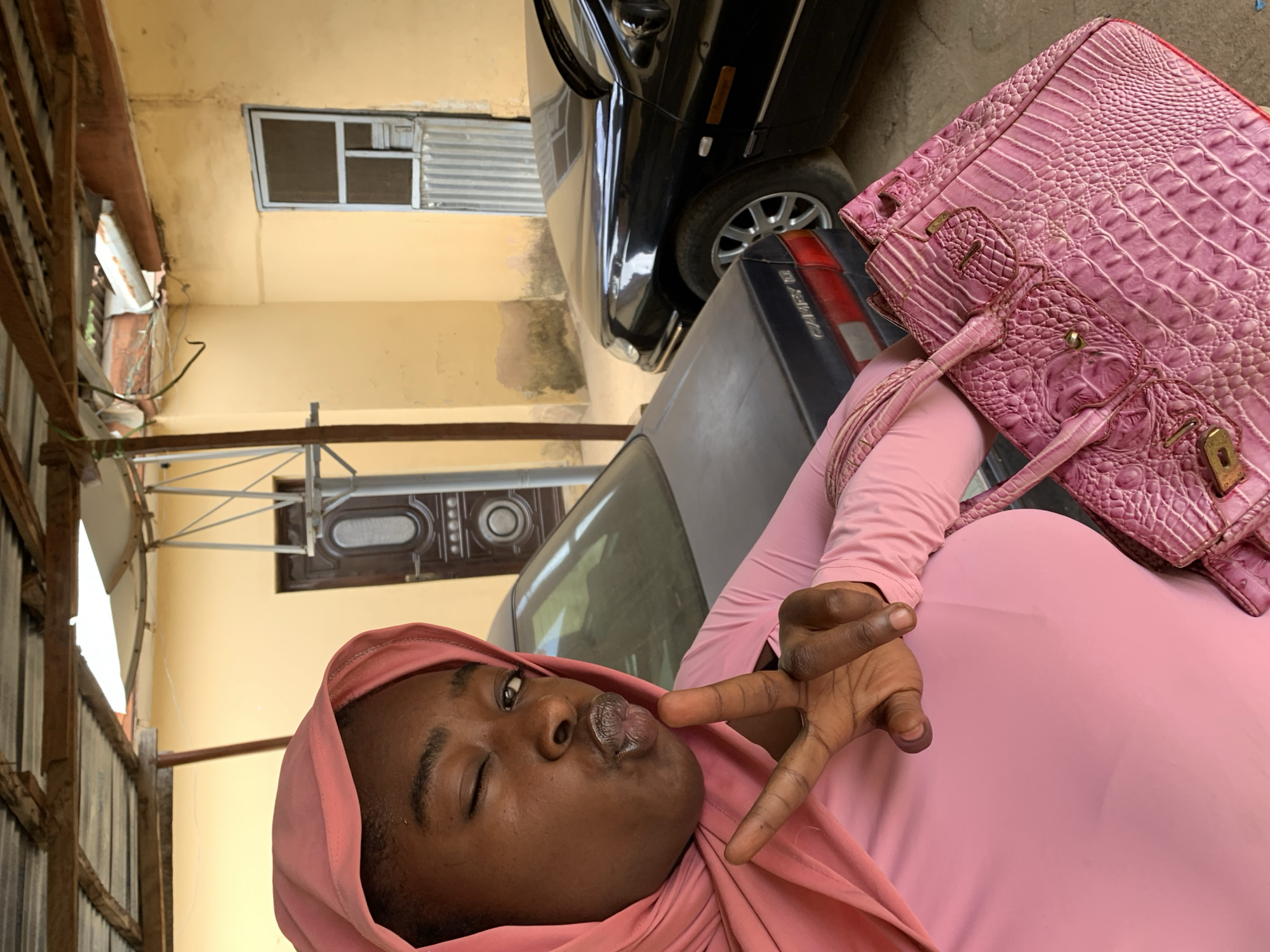 The image shows a person with dark skin wearing a pink headscarf and pink clothing, giving a peace sign while pursing their lips. A pink handbag with a reptile skin pattern is positioned next to them. The background includes two cars, a black one and a gray one, parked near a yellow-painted building with a window. The location is Rafin Par, Nigeria.