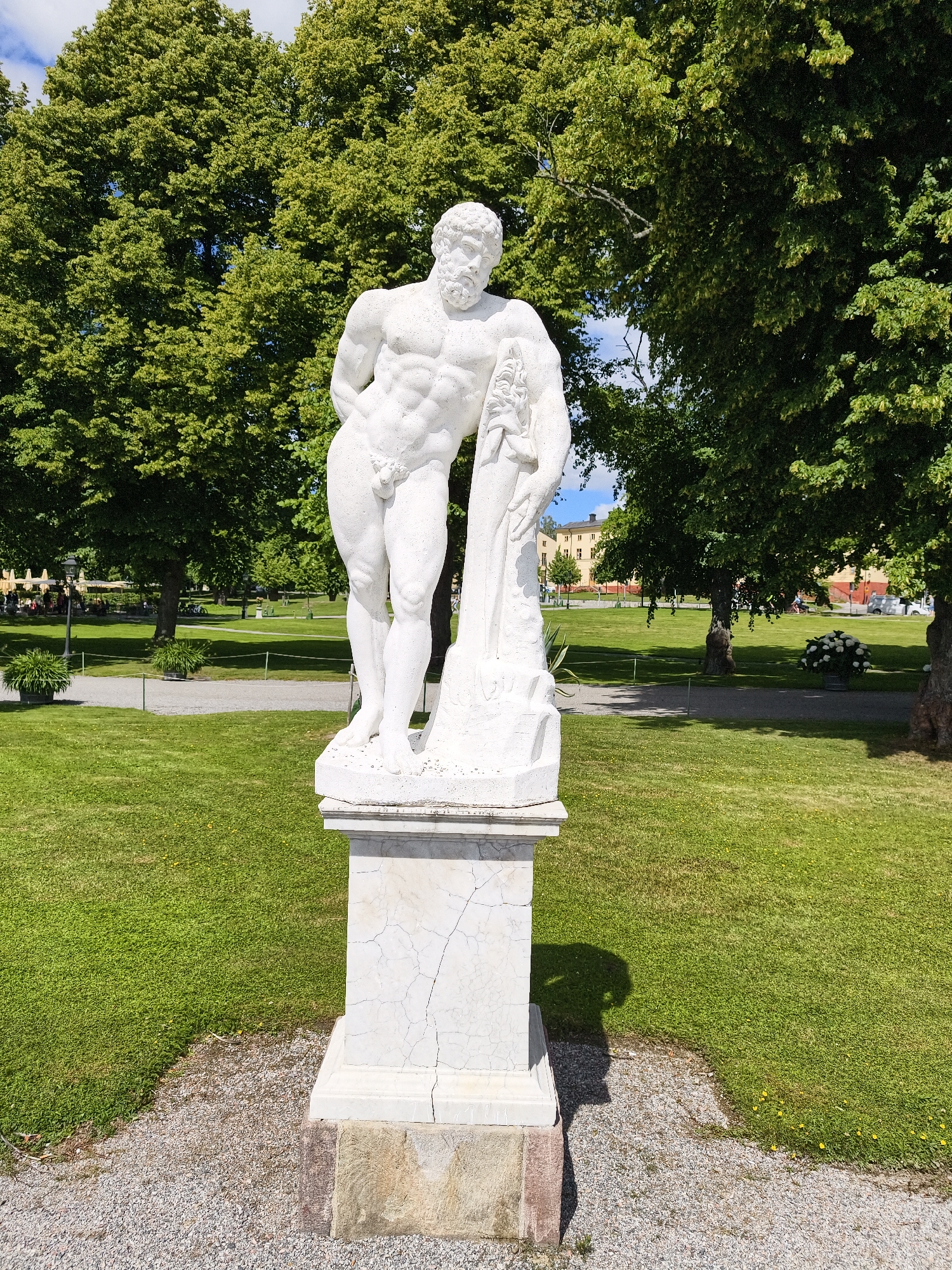 In Ekerö kommun, Sweden, a white marble statue of a muscular, nude male figure stands on a cracked marble pedestal. The statue is positioned on a gravel path in front of a lush green lawn, with large, mature trees providing a dense canopy overhead. In the background, beyond the trees, several yellow buildings are visible, and a few people are seated at tables under umbrellas on the grass. The sky is a bright blue with scattered clouds, suggesting a sunny day. The overall scene depicts a public park or garden setting with a classical sculpture as a focal point.
