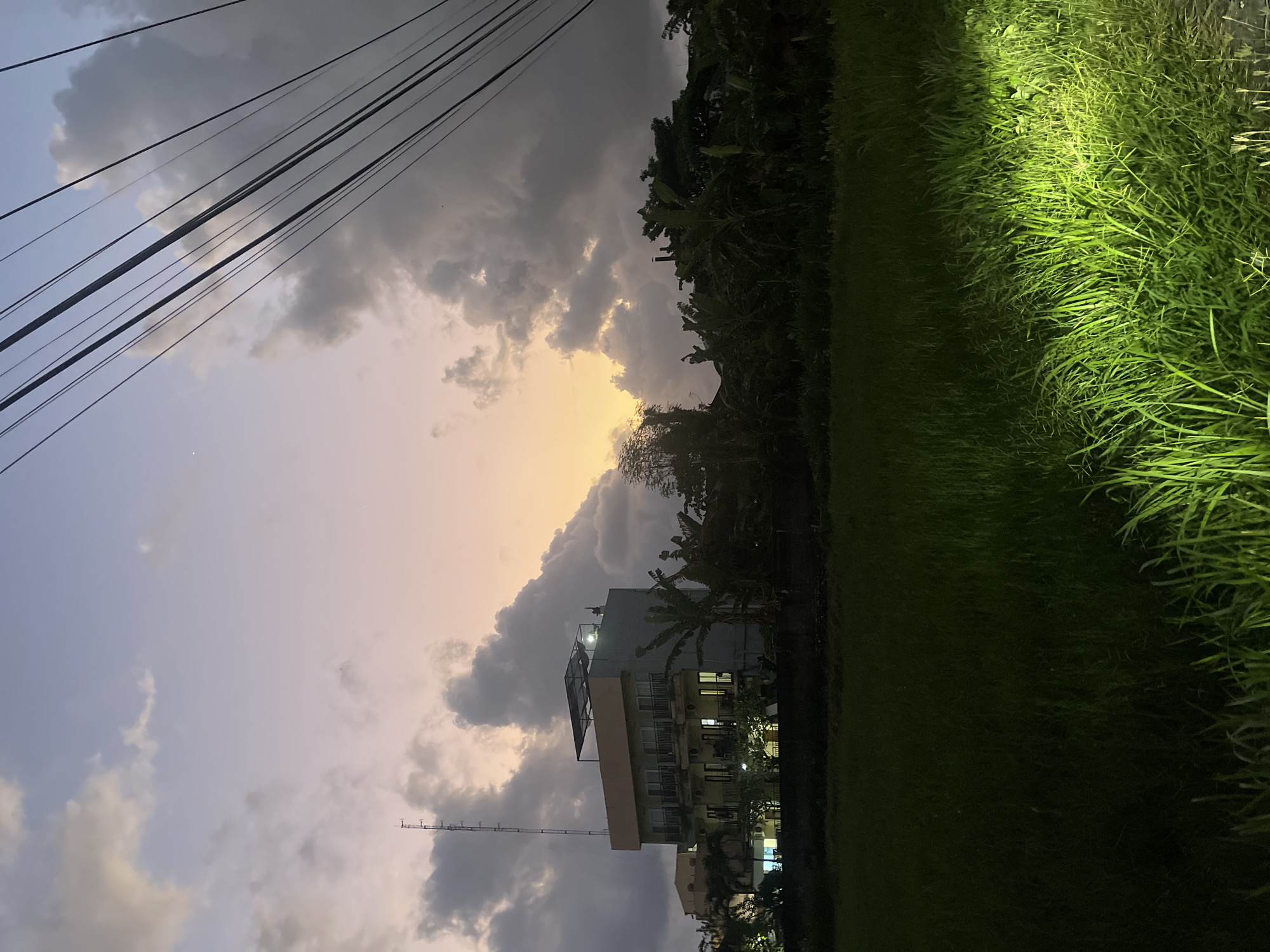 The image depicts a scene at dusk in Kuta, Indonesia, with a building and vegetation in the foreground. The sky displays a gradient of colors, transitioning from a pale blue at the top to soft oranges and yellows near the horizon where the sun is setting. Dark, voluminous clouds are scattered across the sky, some illuminated by the fading sunlight.
A multi-story building, likely a hotel or apartment complex, stands to the left of the frame. It is grey with light-colored trim and features multiple balconies and windows, some of which are lit from within. A tall communication tower is visible on the roof of this building. In front of the building and stretching across the middle ground, there is dense foliage, including what appear to be banana trees and other tropical plants.
The lower portion of the image is dominated by a field of lush green grass, possibly a rice paddy, which is partially illuminated by a bright, artificial light source from the right foreground. This light casts a strong glow on the grass, creating a stark contrast with the darker, unlit areas. Overhead, a cluster of dark power lines stretches diagonally across the upper right portion of the frame, silhouetted against the sky. The overall atmosphere is tranquil, capturing the transition from day to night in a tropical setting.